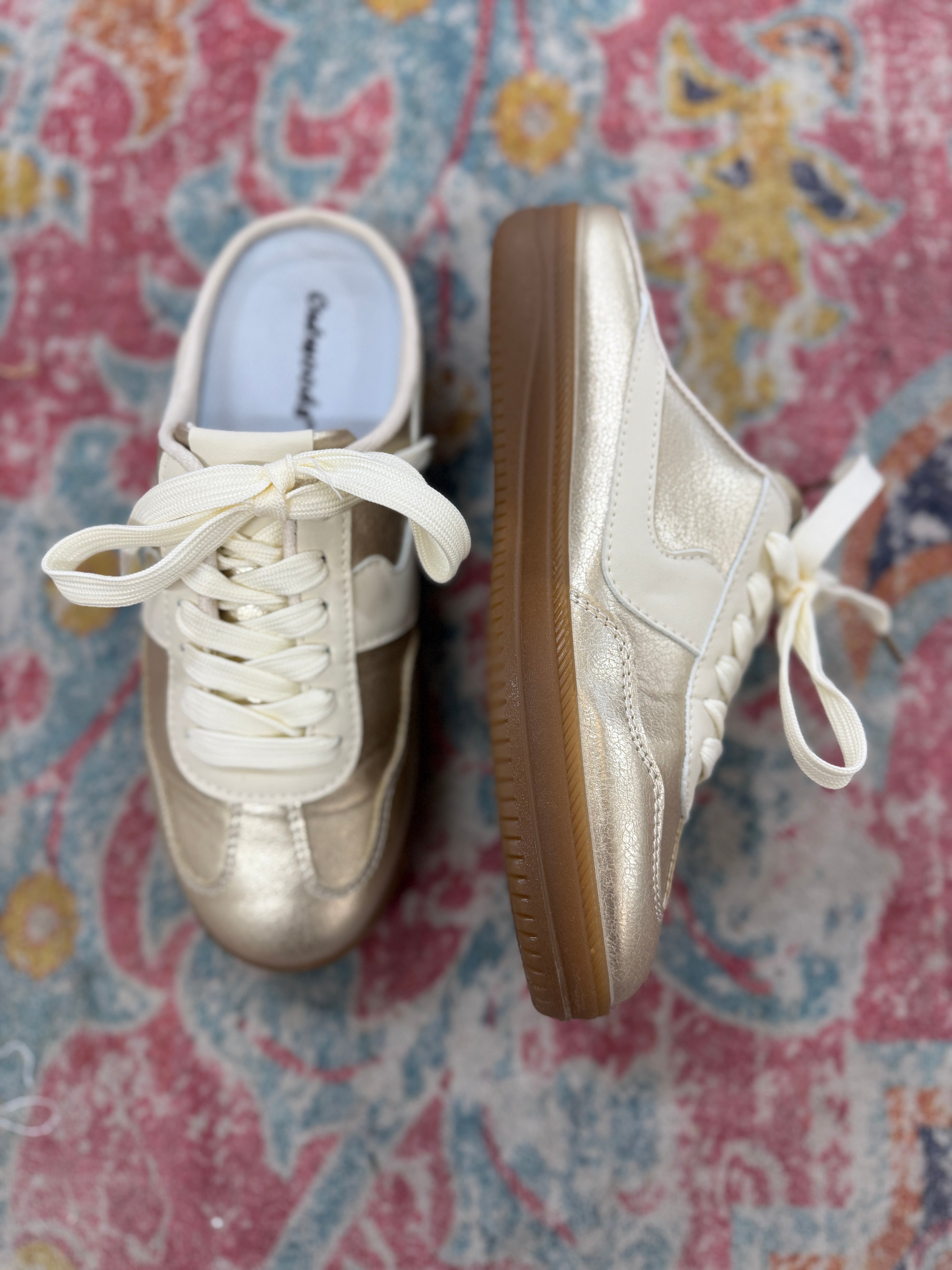 Gold metallic slip-on sneakers with white laces and gum soles on colorful patterned rug
