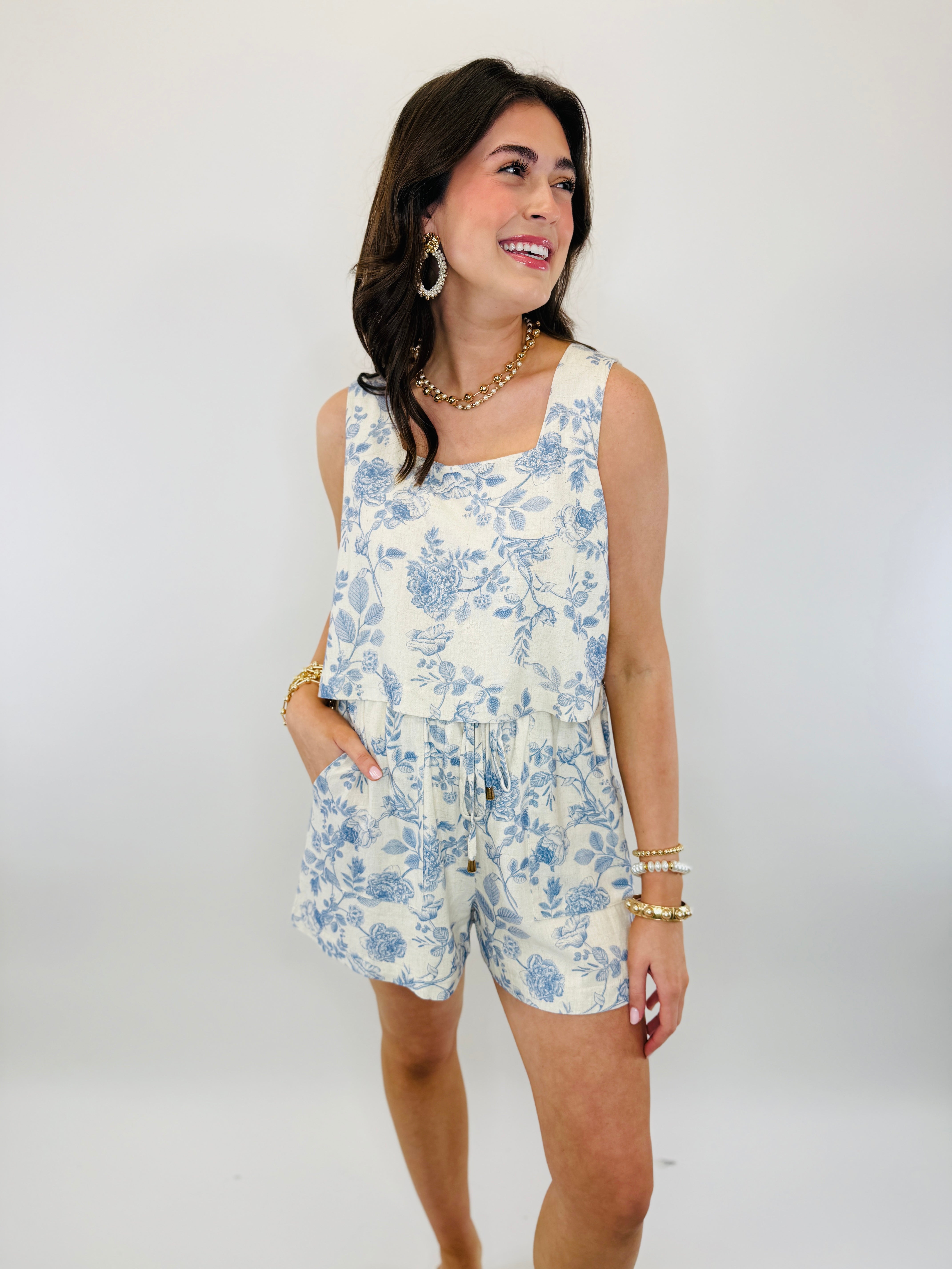 Woman wearing blue and white floral sleeveless romper with shorts, styled with gold jewelry and bracelet stack.