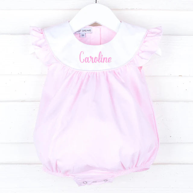 Pink baby bubble romper with white collar and "Caroline" embroidered text, displayed on white wooden background.