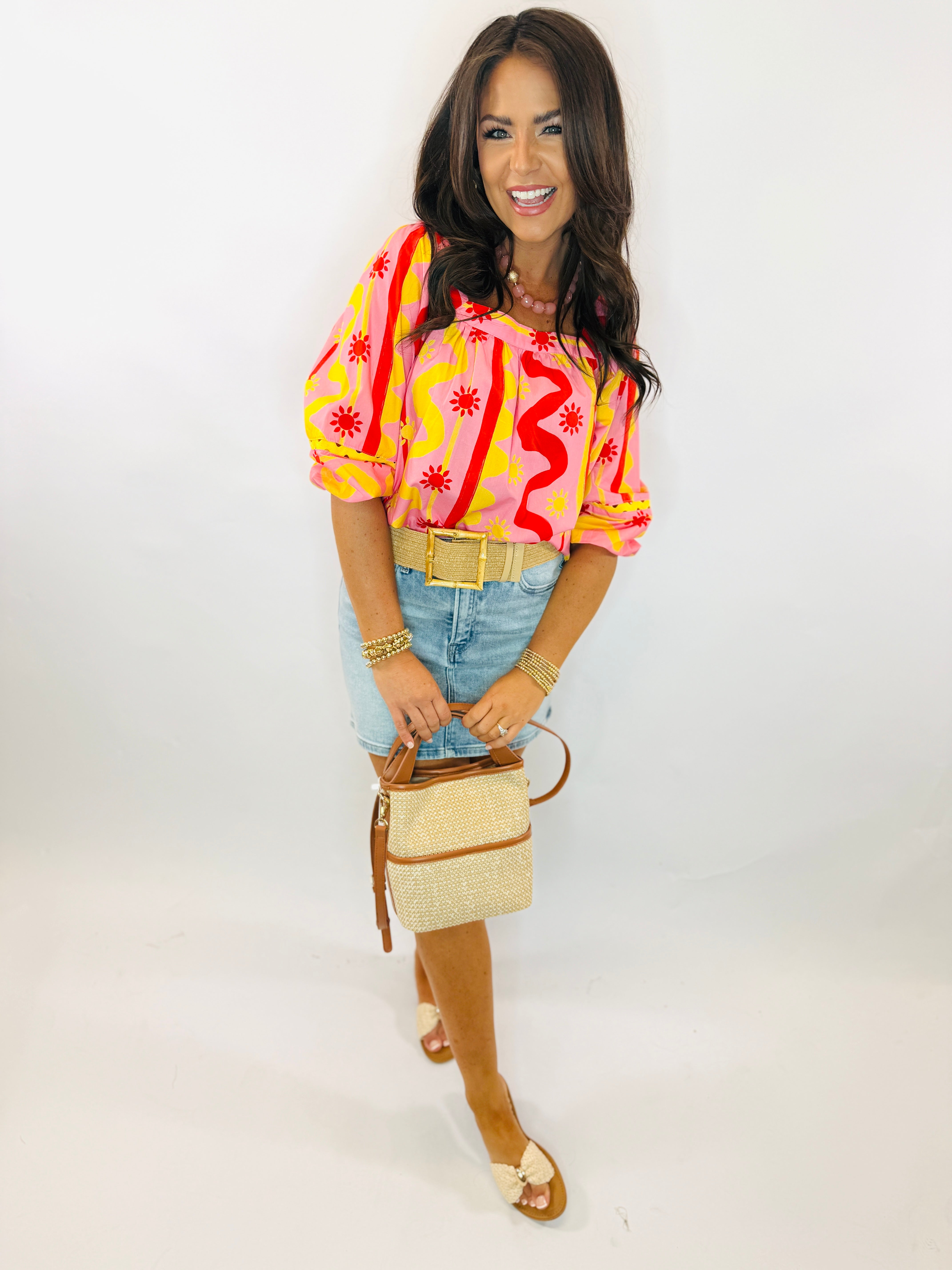 Woman wearing colorful Emmie Top with pink, yellow, and red swirl pattern, paired with denim skirt and woven handbag.