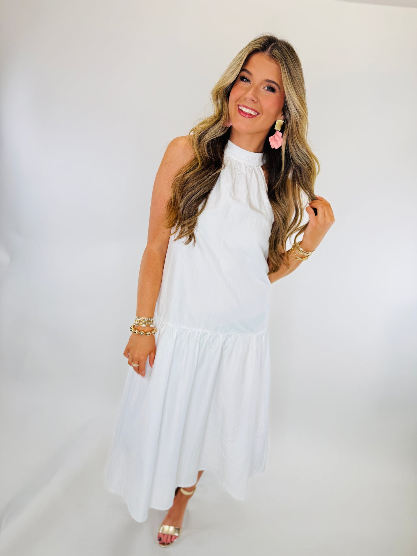 Woman wearing white sleeveless halter dress with tiered ruffle hem, pink tassel earrings, and gold bracelets