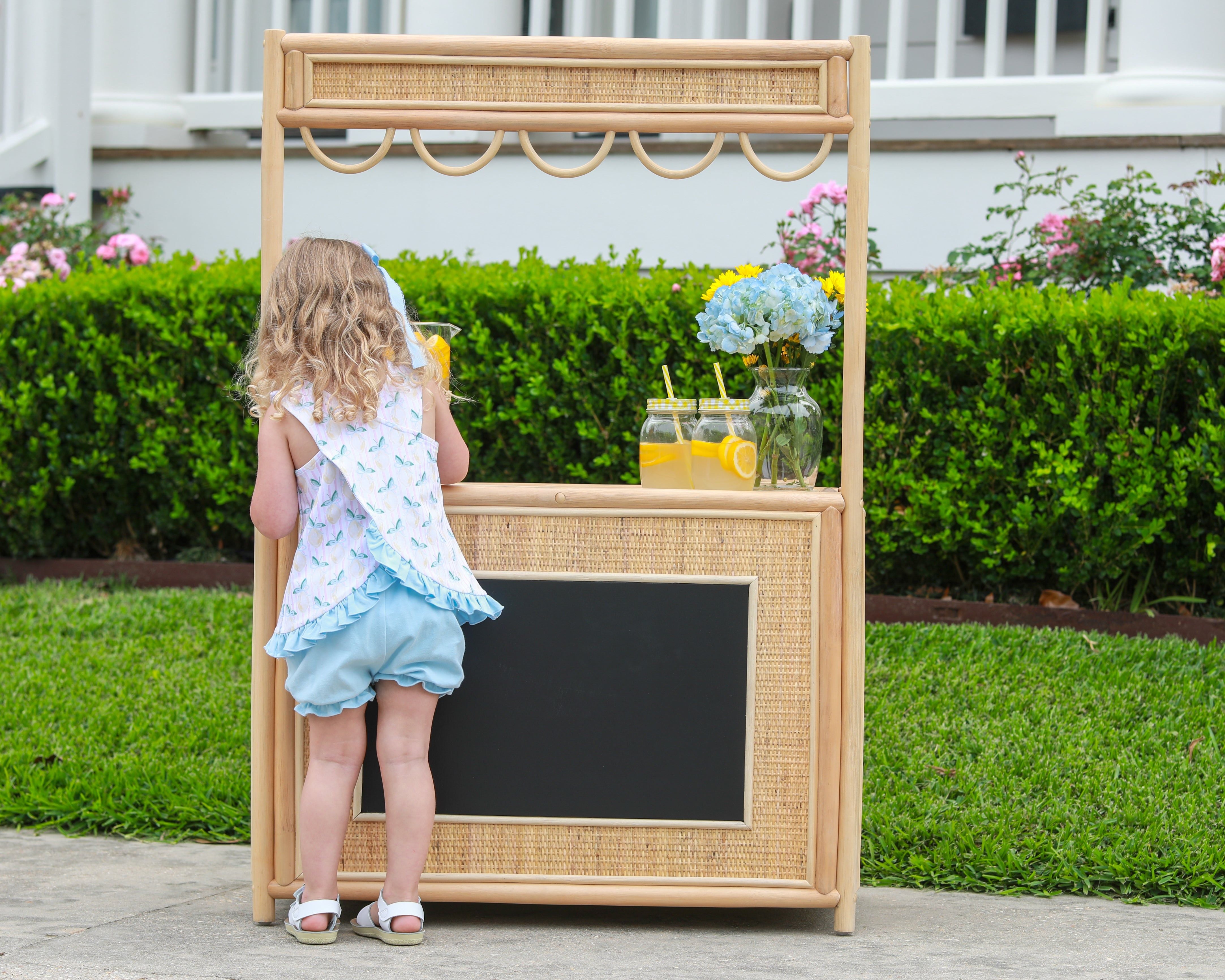 Young child in light blue ruffled top and bloomer shorts standing at wooden lemonade stand with chalkboard sign.