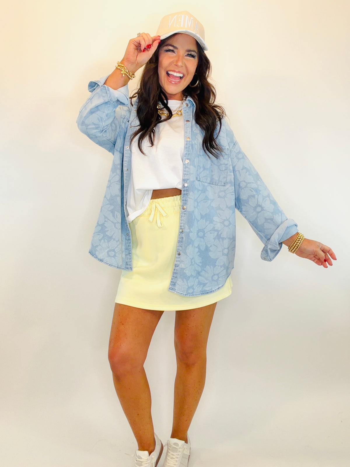 Woman wearing light blue denim shacket over white crop top and yellow mini skirt with white sneakers and cap