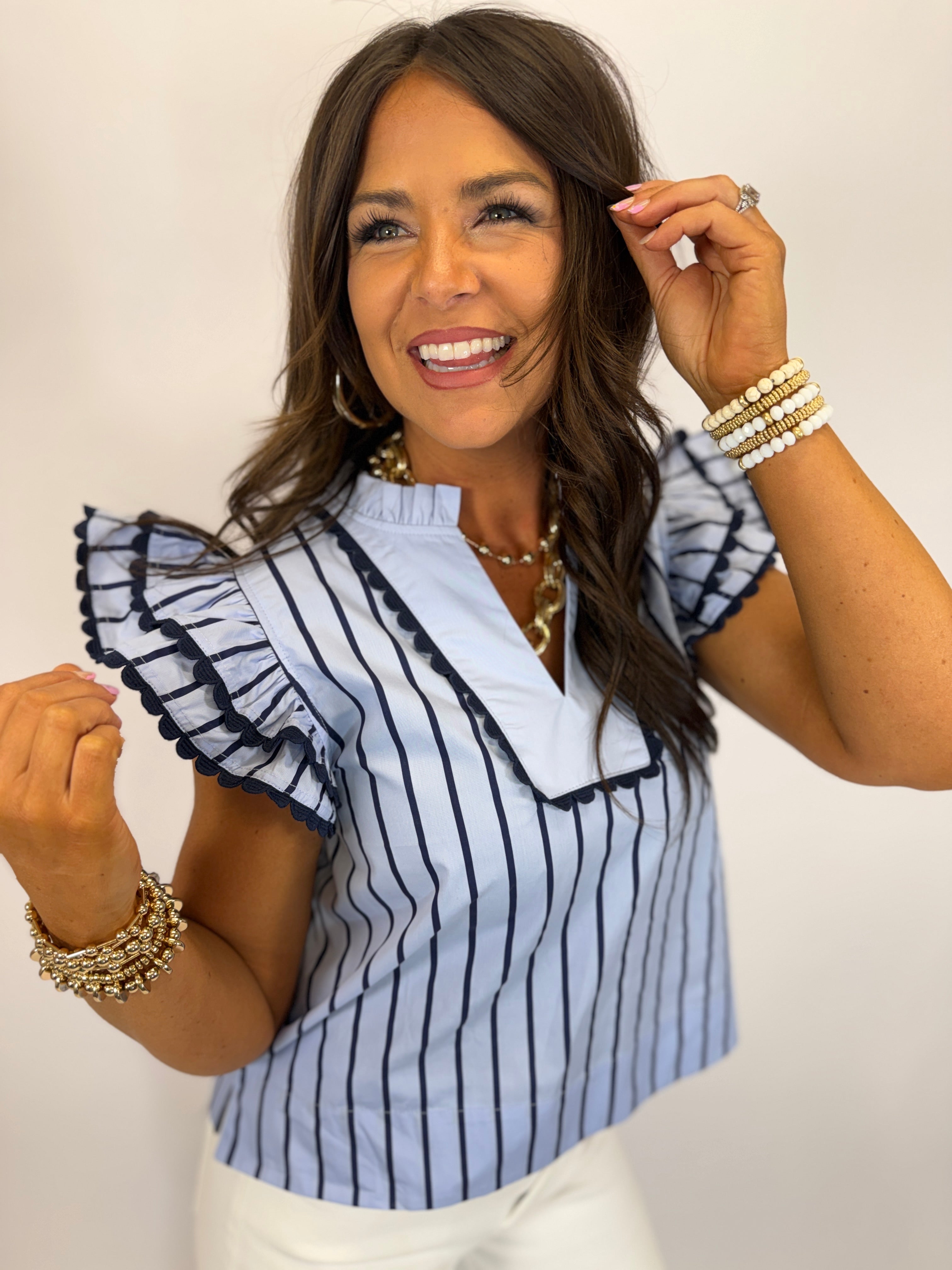 Woman wearing white striped ruffle sleeve top with navy trim and gold jewelry, smiling at camera.