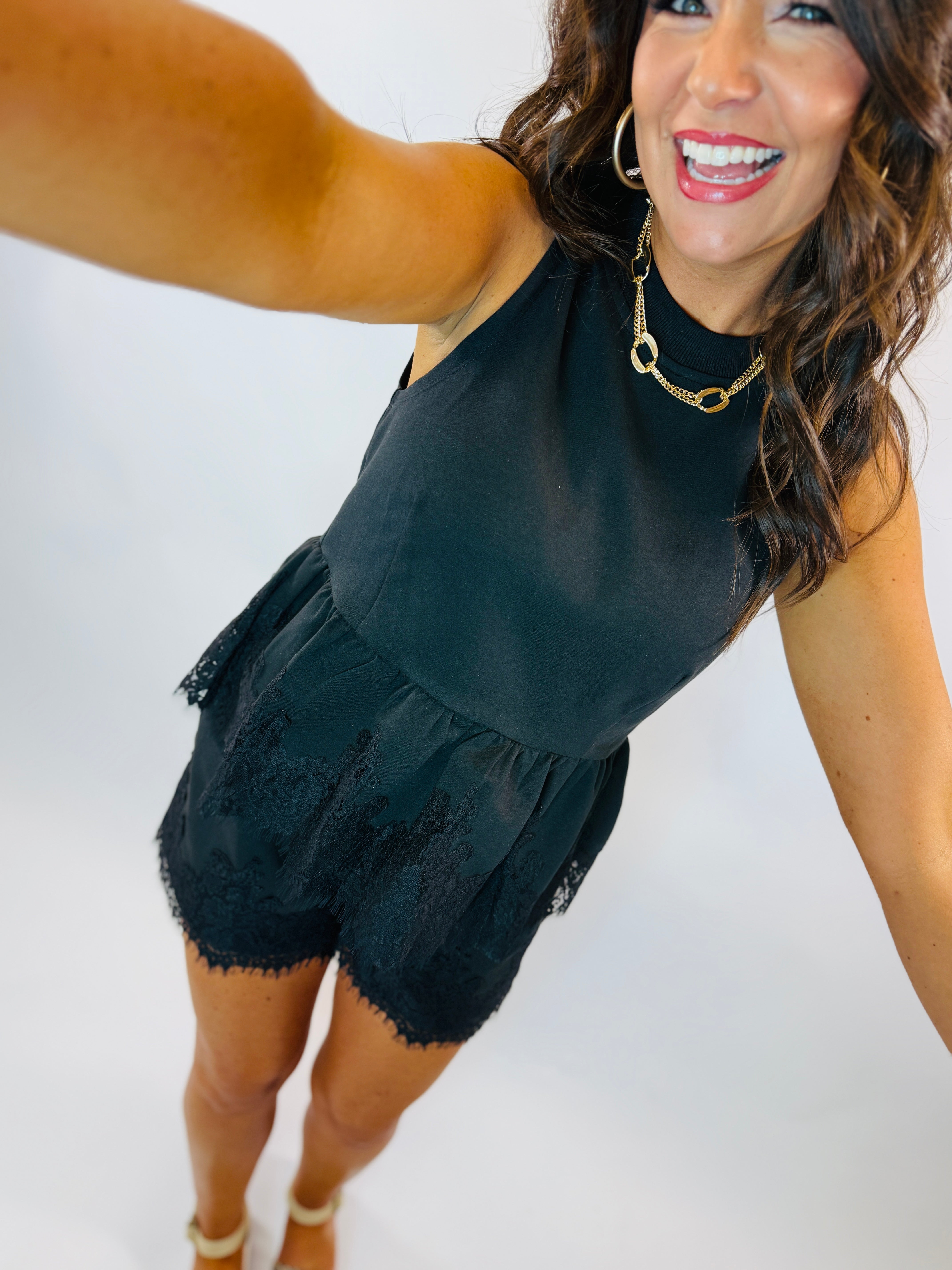 Woman wearing black sleeveless top with lace trim hem and gold chain necklace, smiling and posing.
