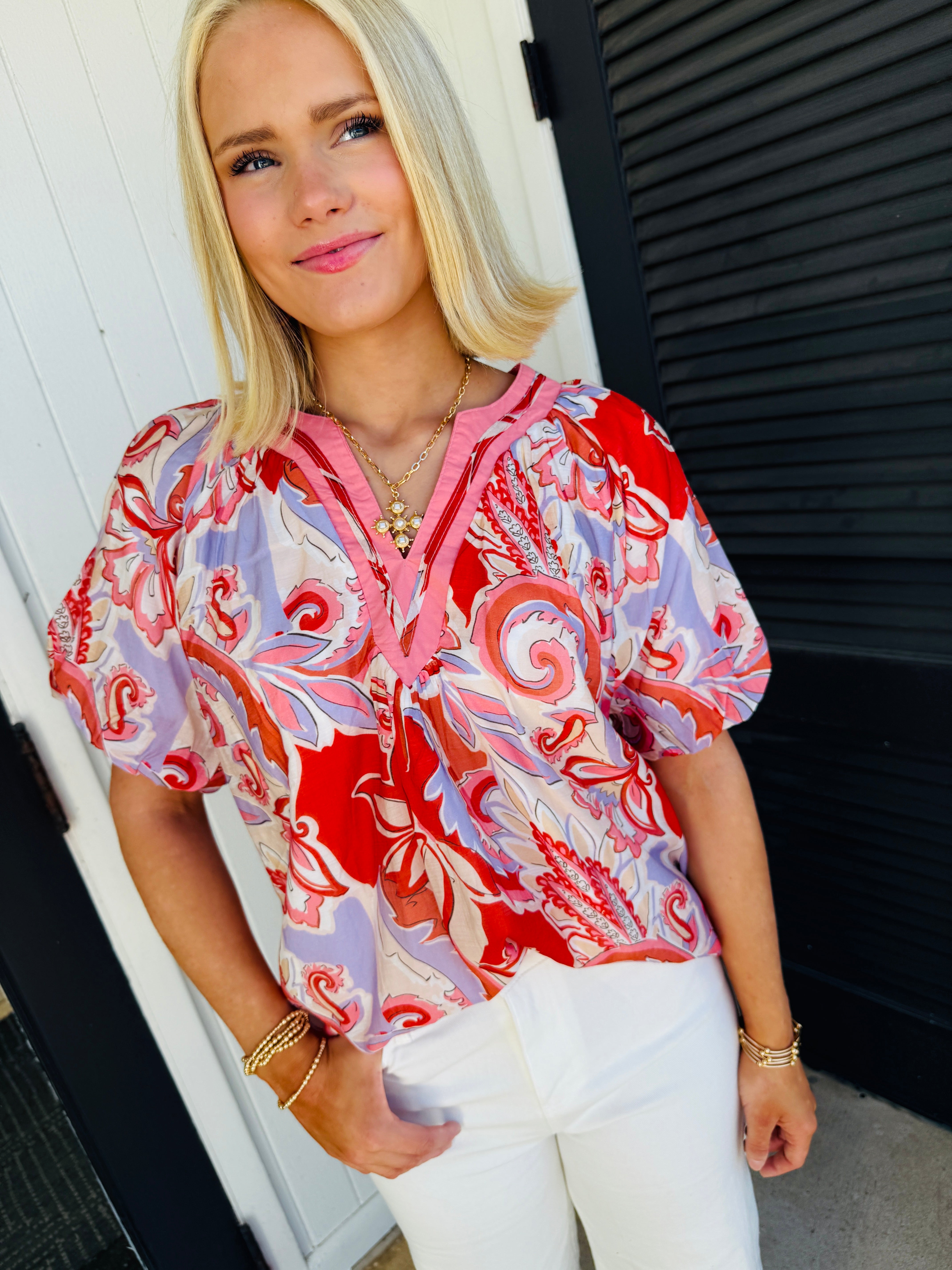 Woman wearing colorful patterned puff sleeve blouse with pink trim and white pants, styled with gold jewelry.