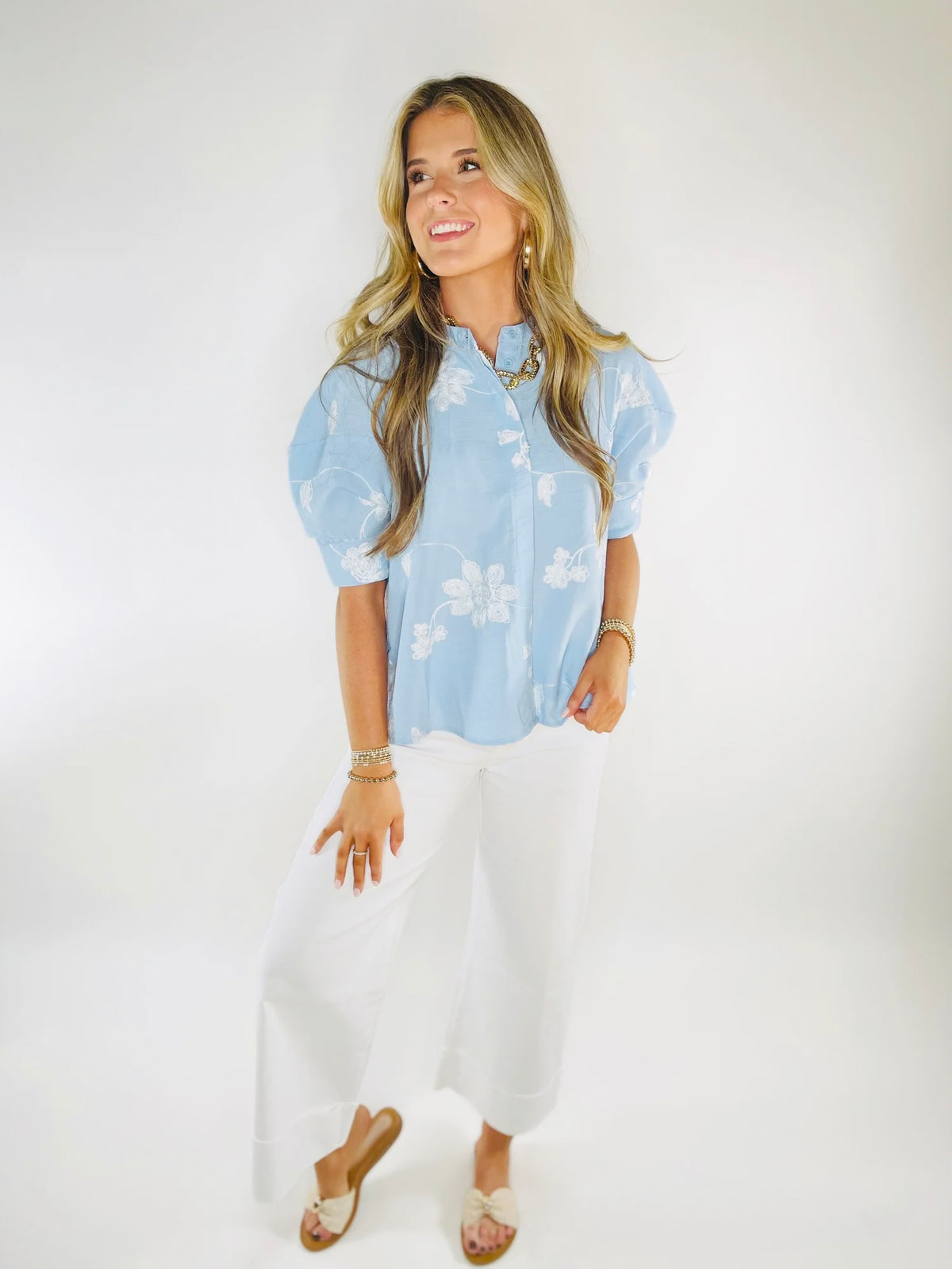Woman wearing light blue floral short-sleeve button-up blouse with white pants and sandals, smiling while looking away