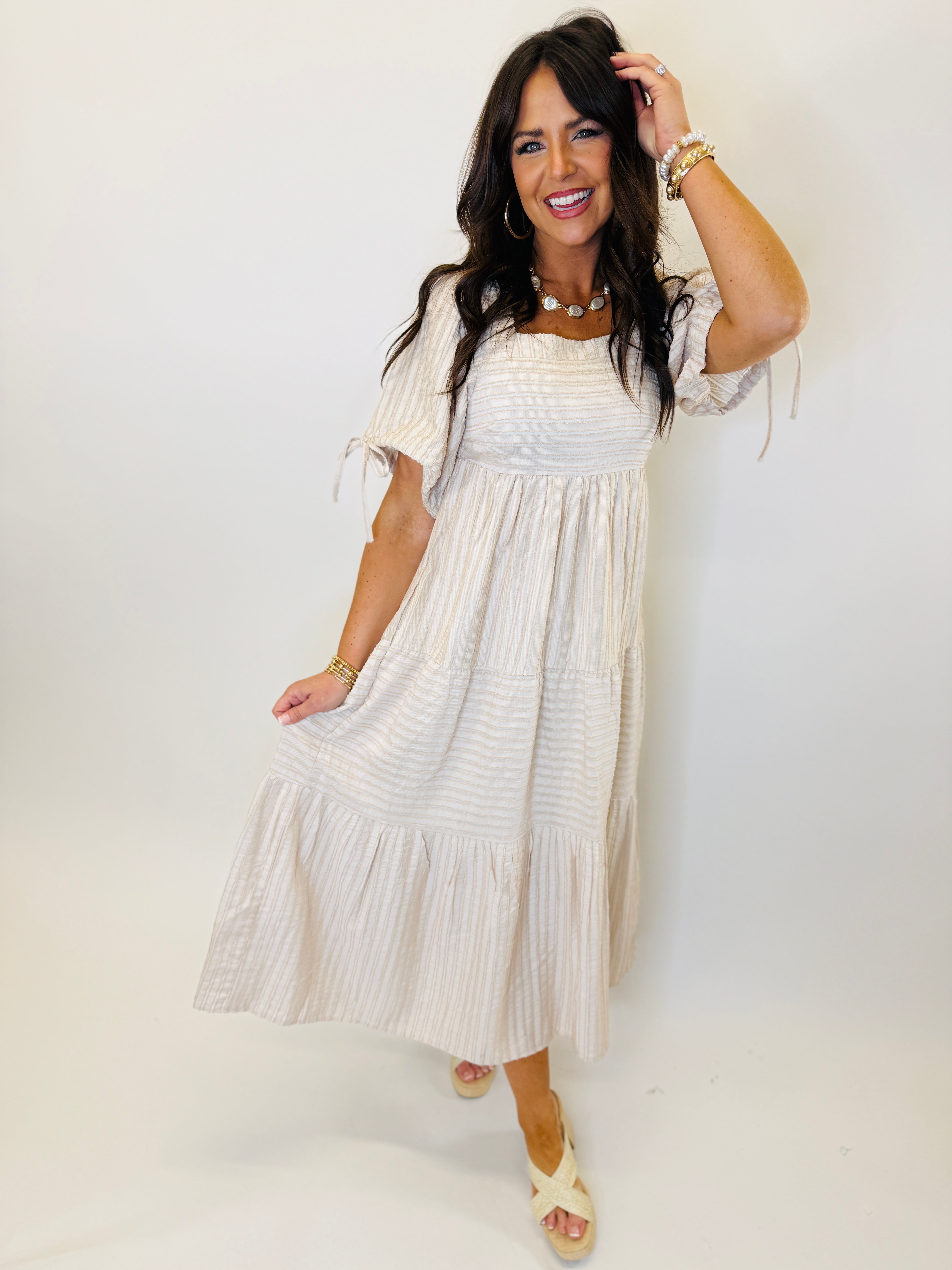Woman wearing taupe striped tiered maxi dress with off-shoulder sleeves and tie details, styled with gold jewelry.