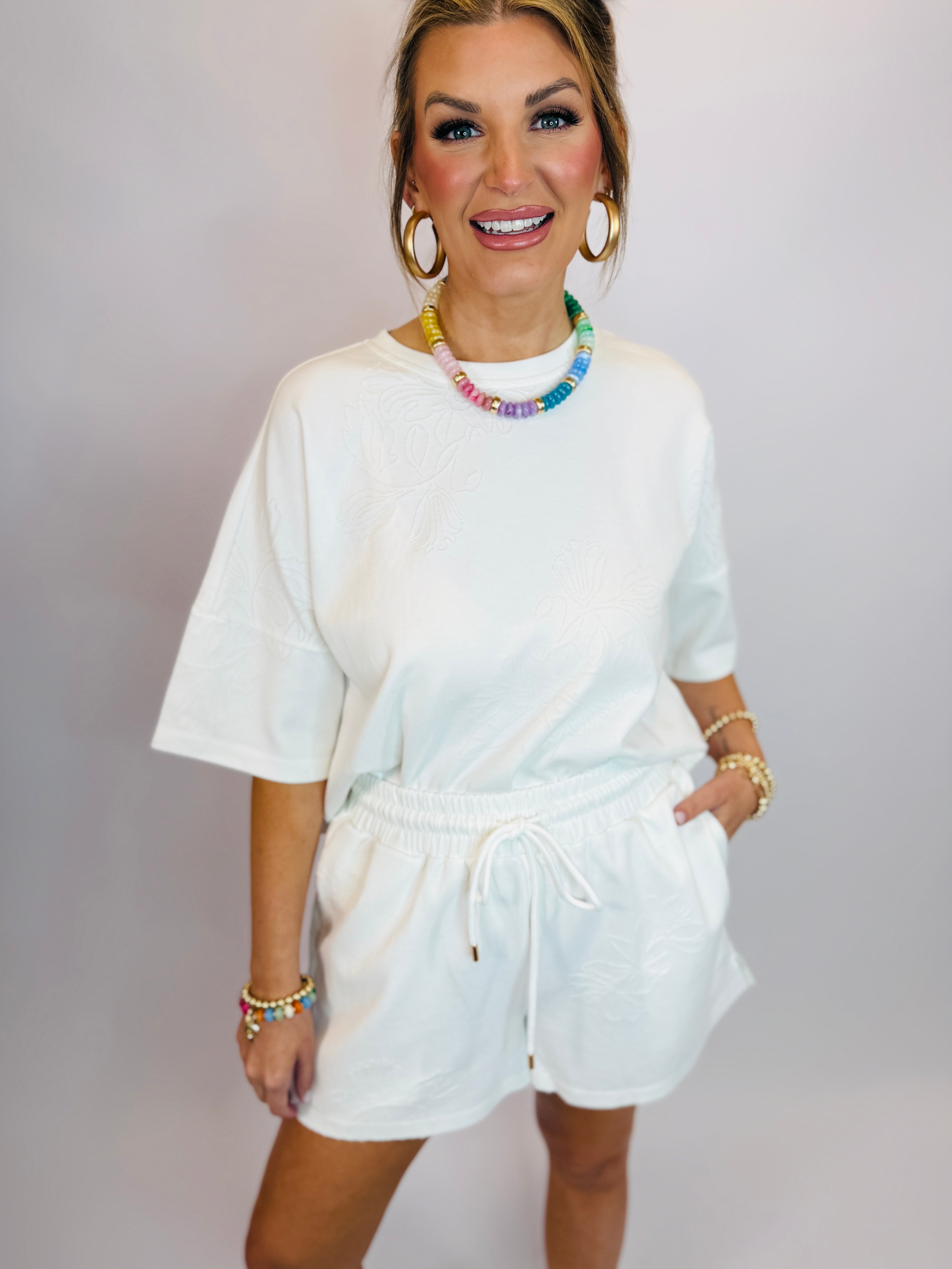 Woman wearing white oversized short-sleeve top with drawstring waistband, styled with colorful beaded necklace and hoop ea...