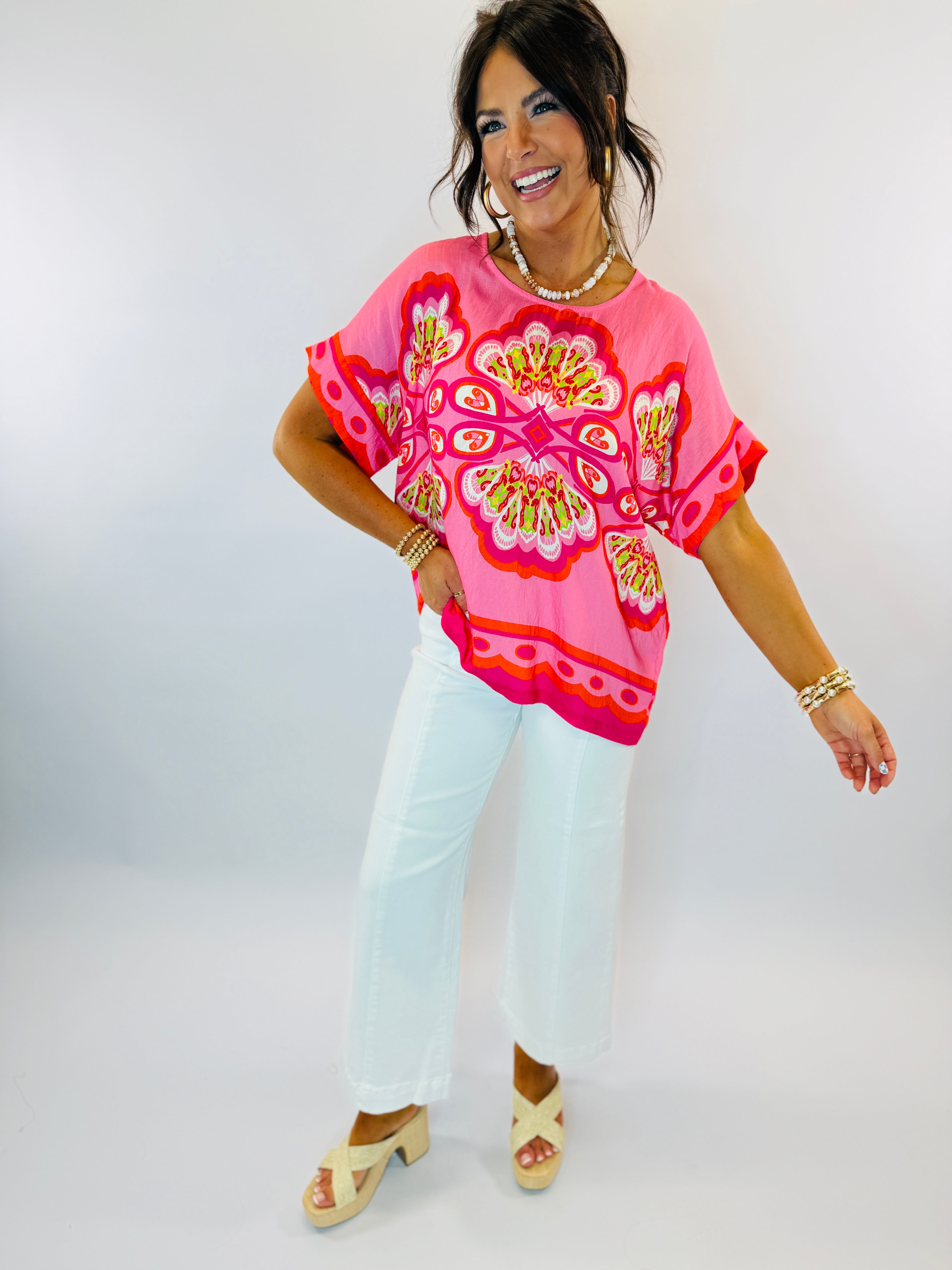 Woman wearing bright pink floral embroidered Laci top with white pants and pearl accessories, smiling pose