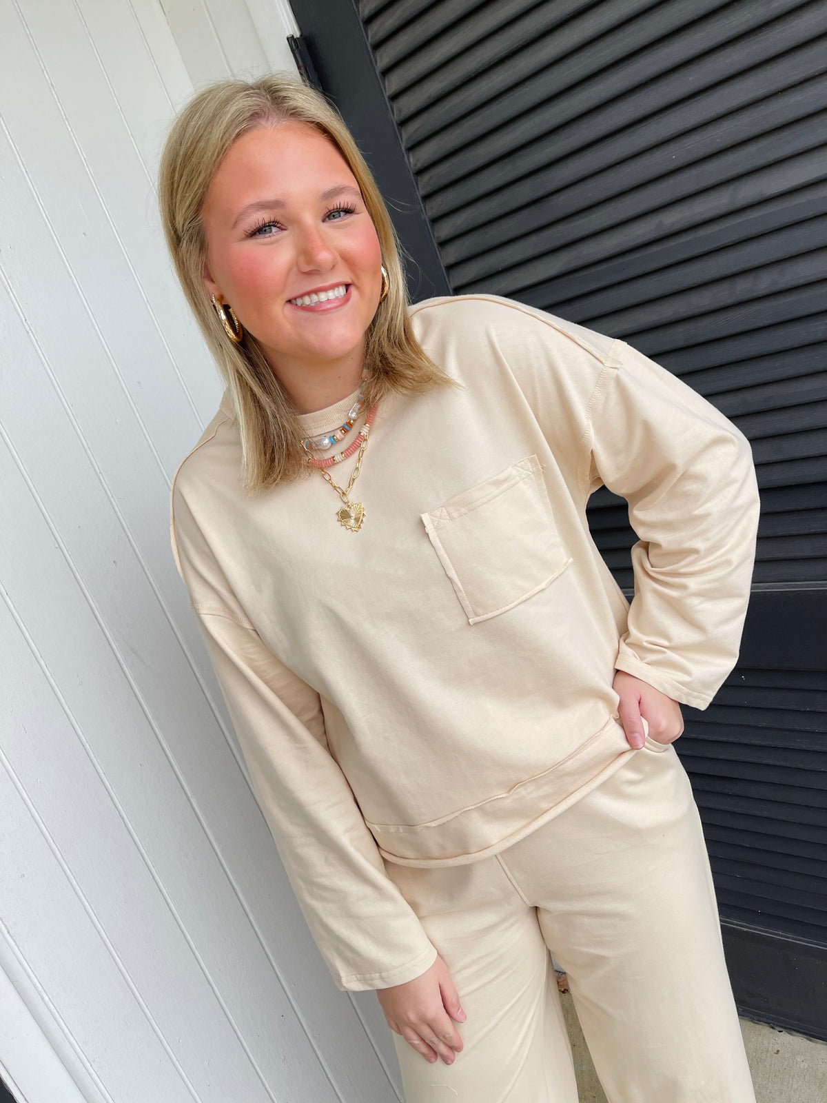 Woman wearing beige oversized sweatshirt with matching sweatpants, styled with layered necklaces and gold hoop earrings.