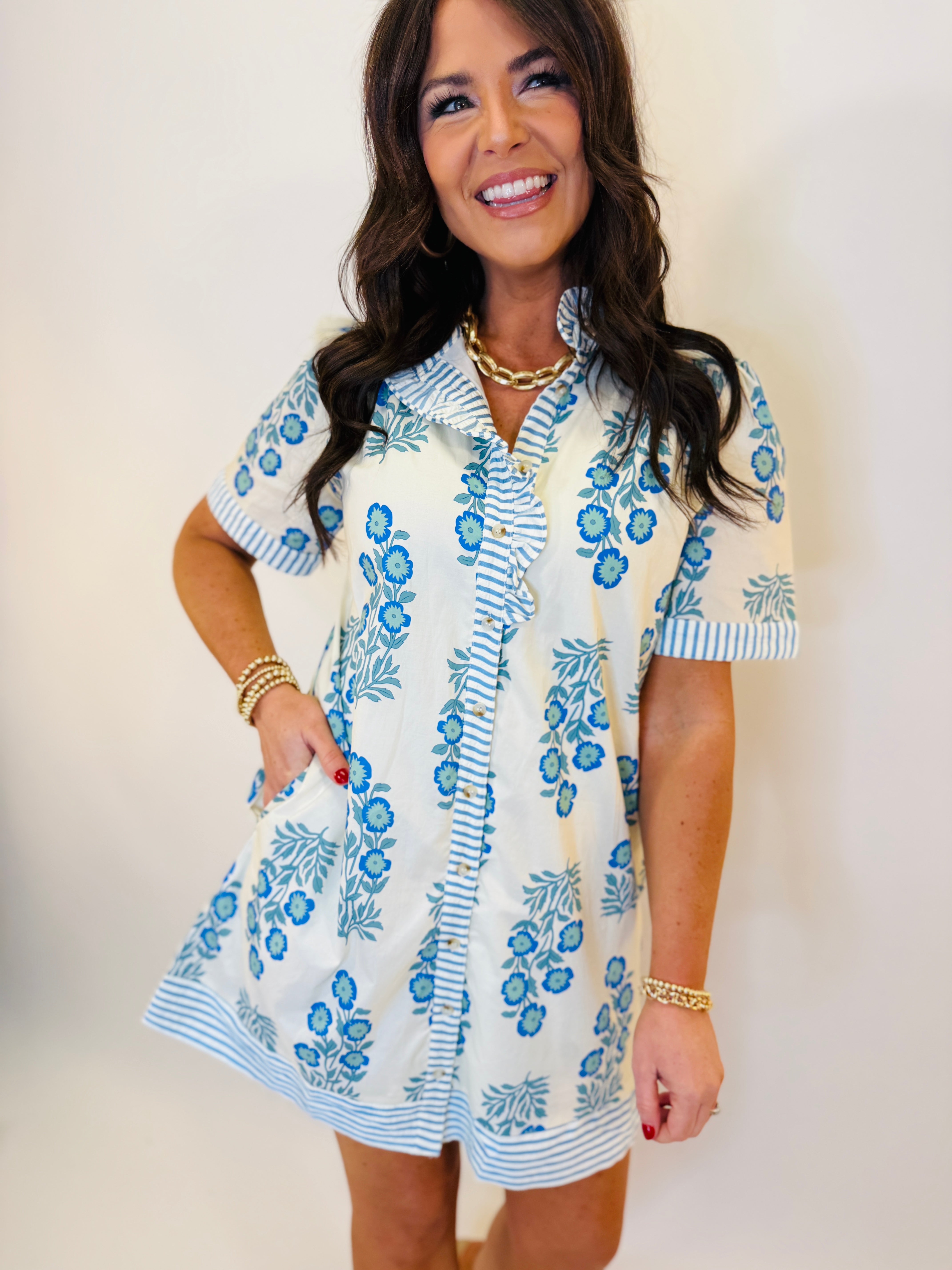 Woman wearing white short-sleeve dress with blue floral pattern and striped trim, styled with gold jewelry.