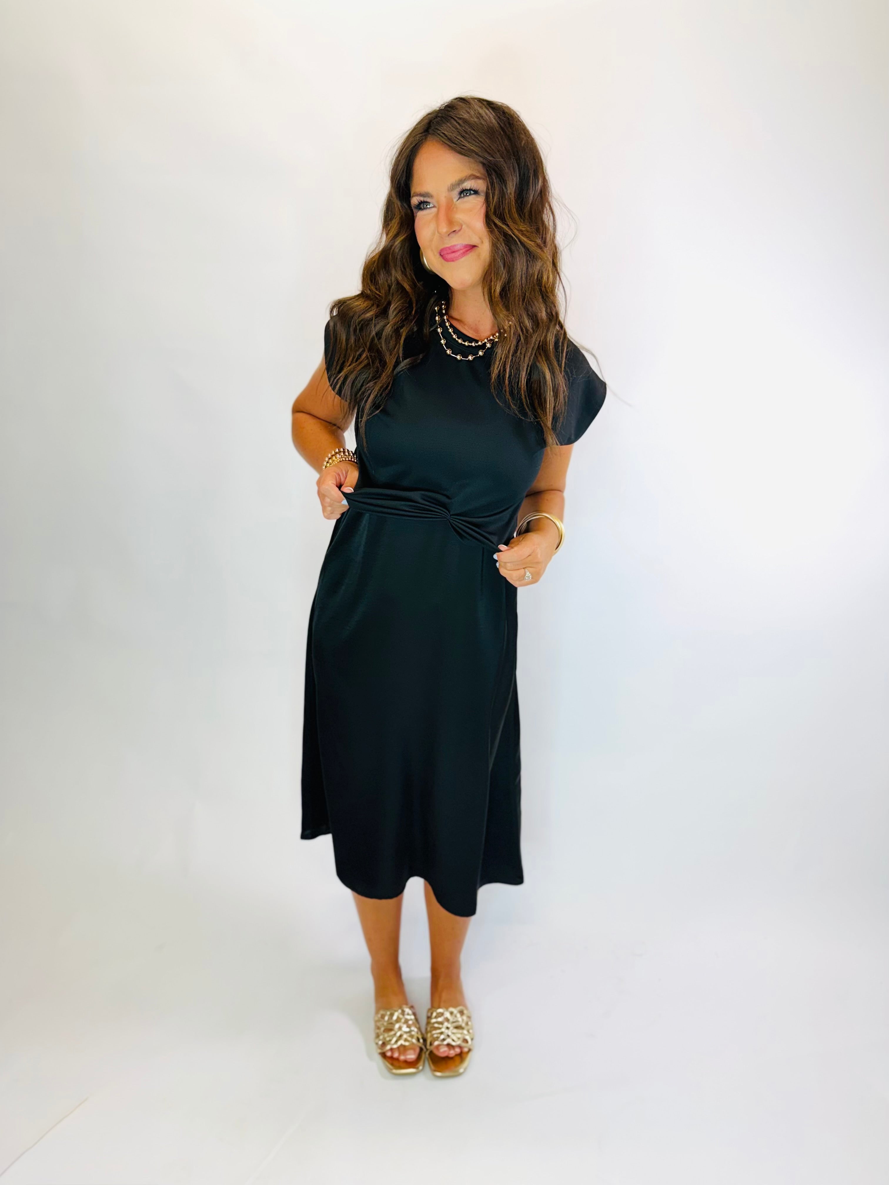 Woman wearing a black midi dress with cap sleeves, twist waist detail, and gold sandals.