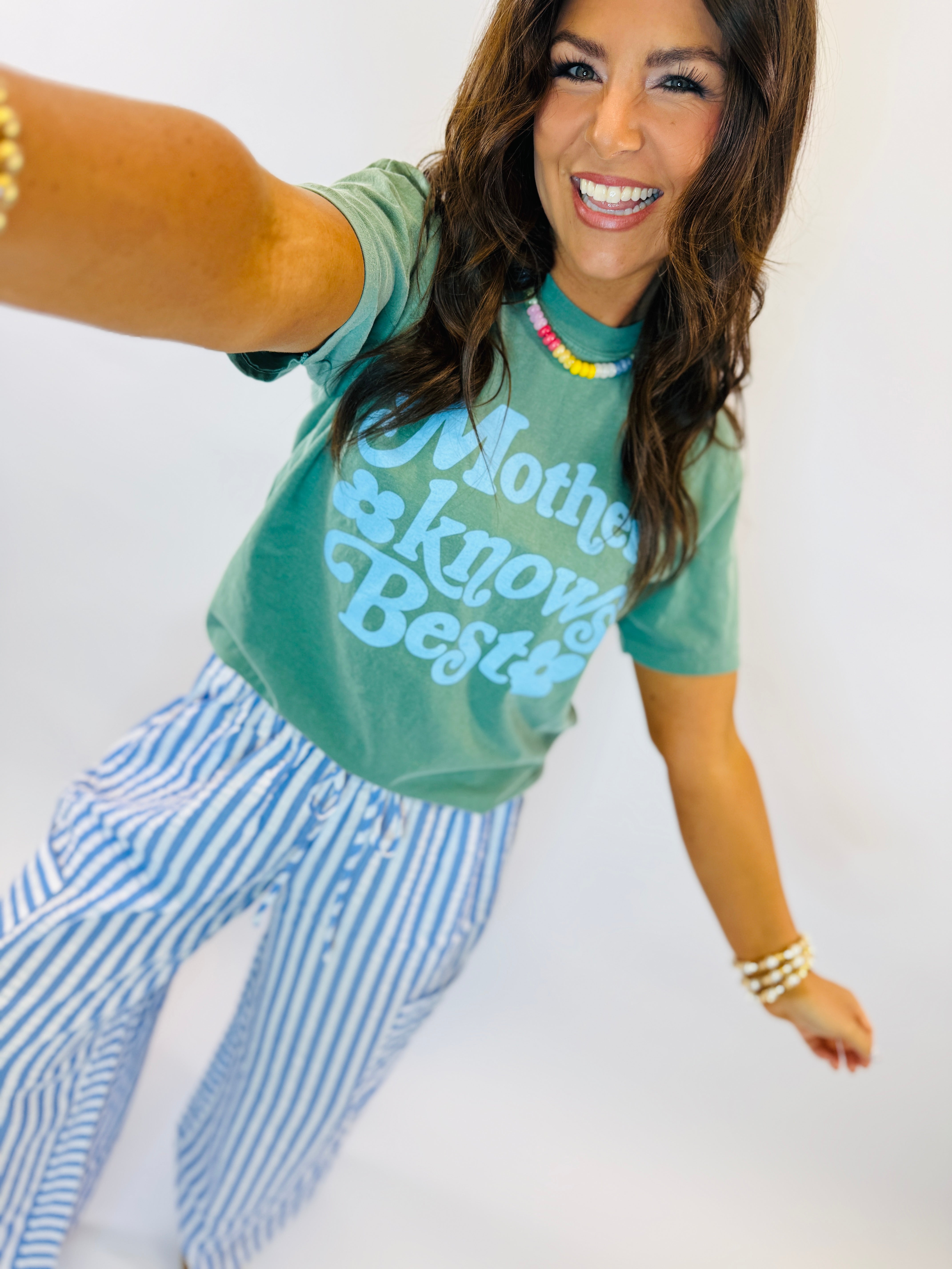 Woman wearing green graphic tee and blue striped Miley pants, posing with colorful beaded necklace.
