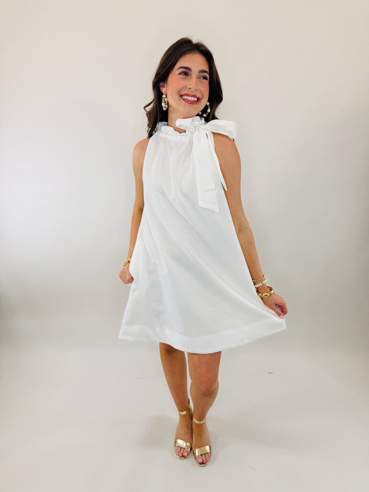 Woman wearing white sleeveless bow-tie neck dress with gold accessories and heeled sandals