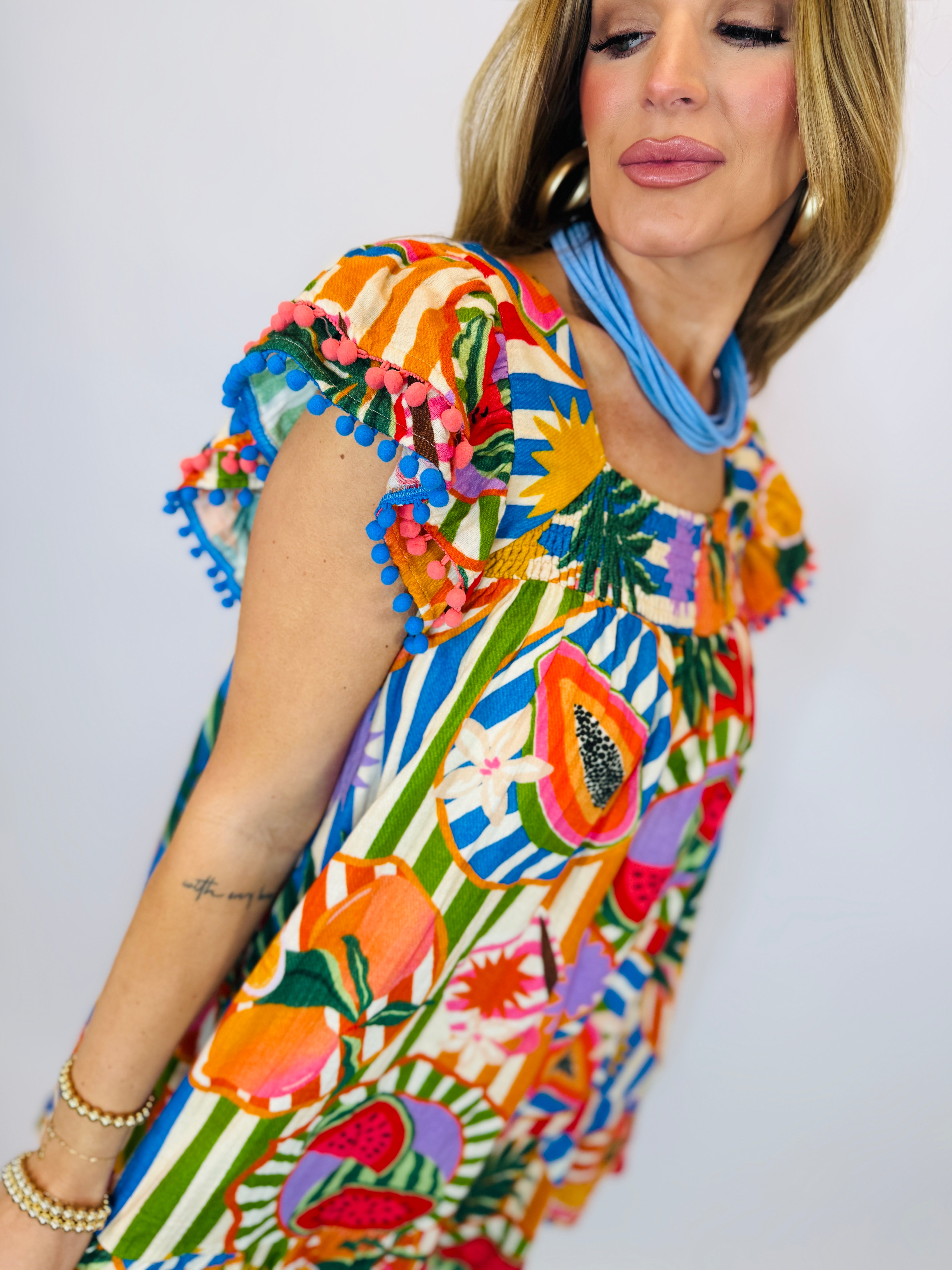 Colorful tropical print Luca dress with pom-pom trim sleeves and vibrant fruit and floral patterns.
