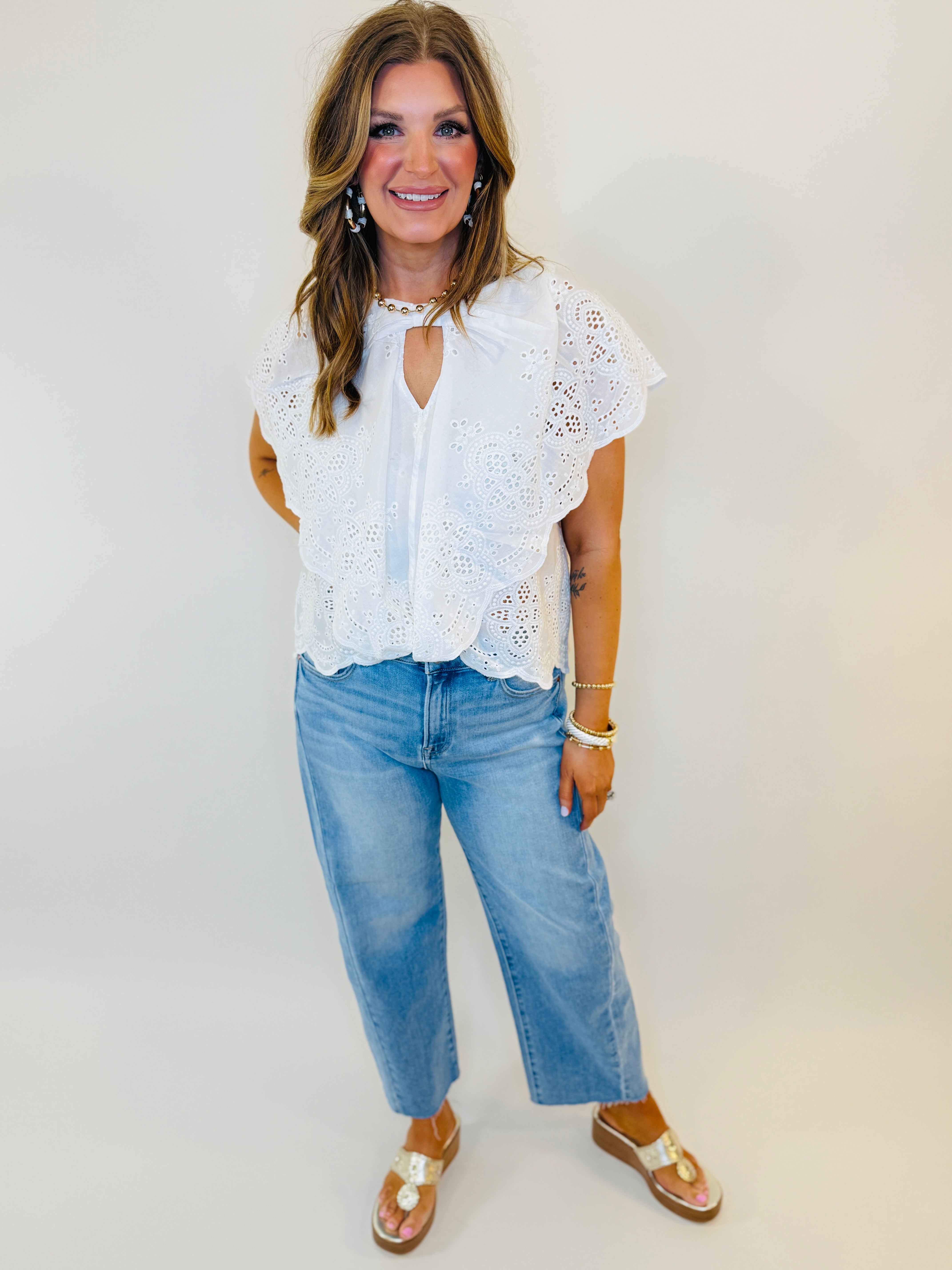 Woman wearing white eyelet lace top with ruffled sleeves, keyhole neckline, paired with light blue jeans and sandals.