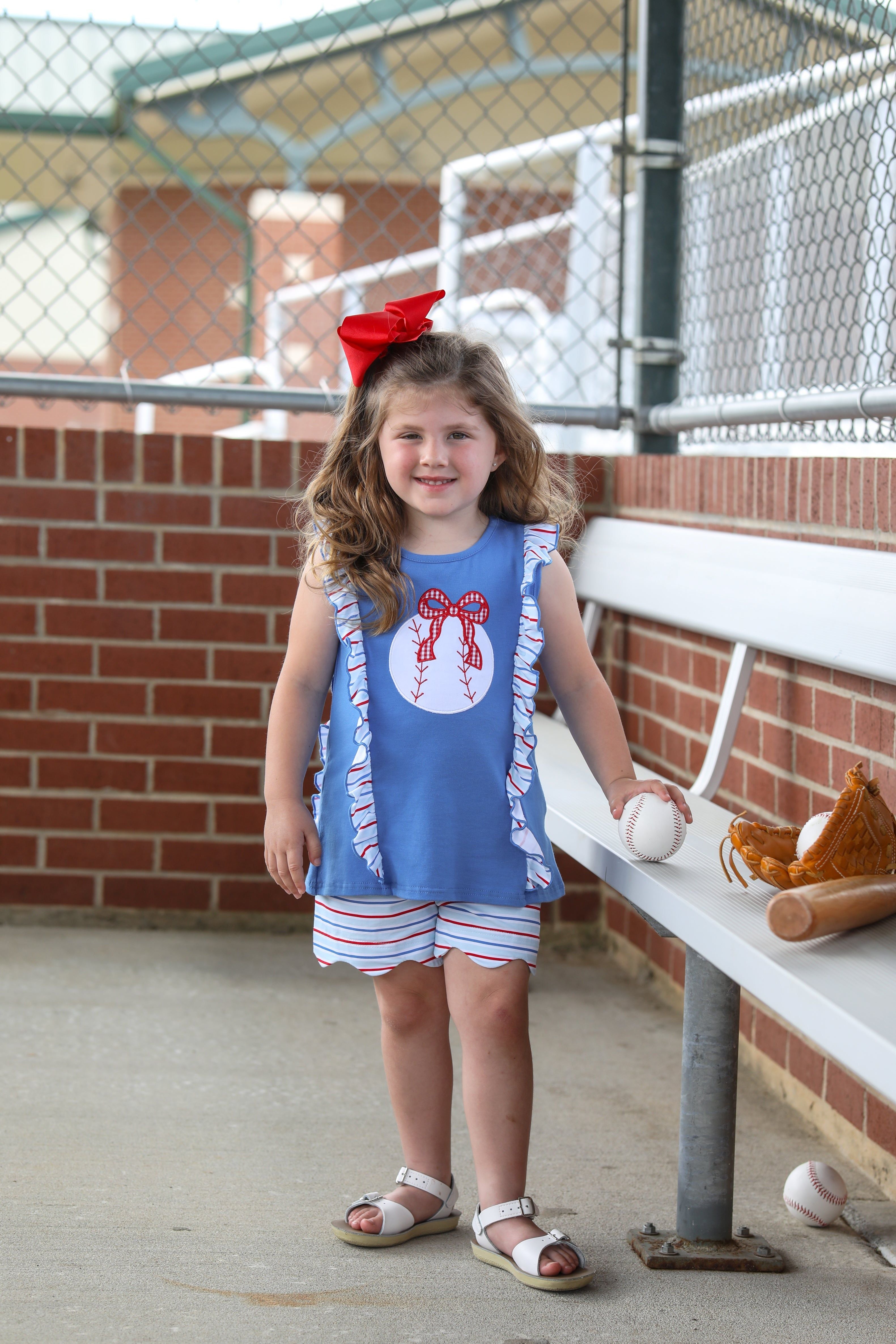 Young girl wearing blue baseball-themed ruffle top and striped shorts set, standing by dugout with baseball equipment.