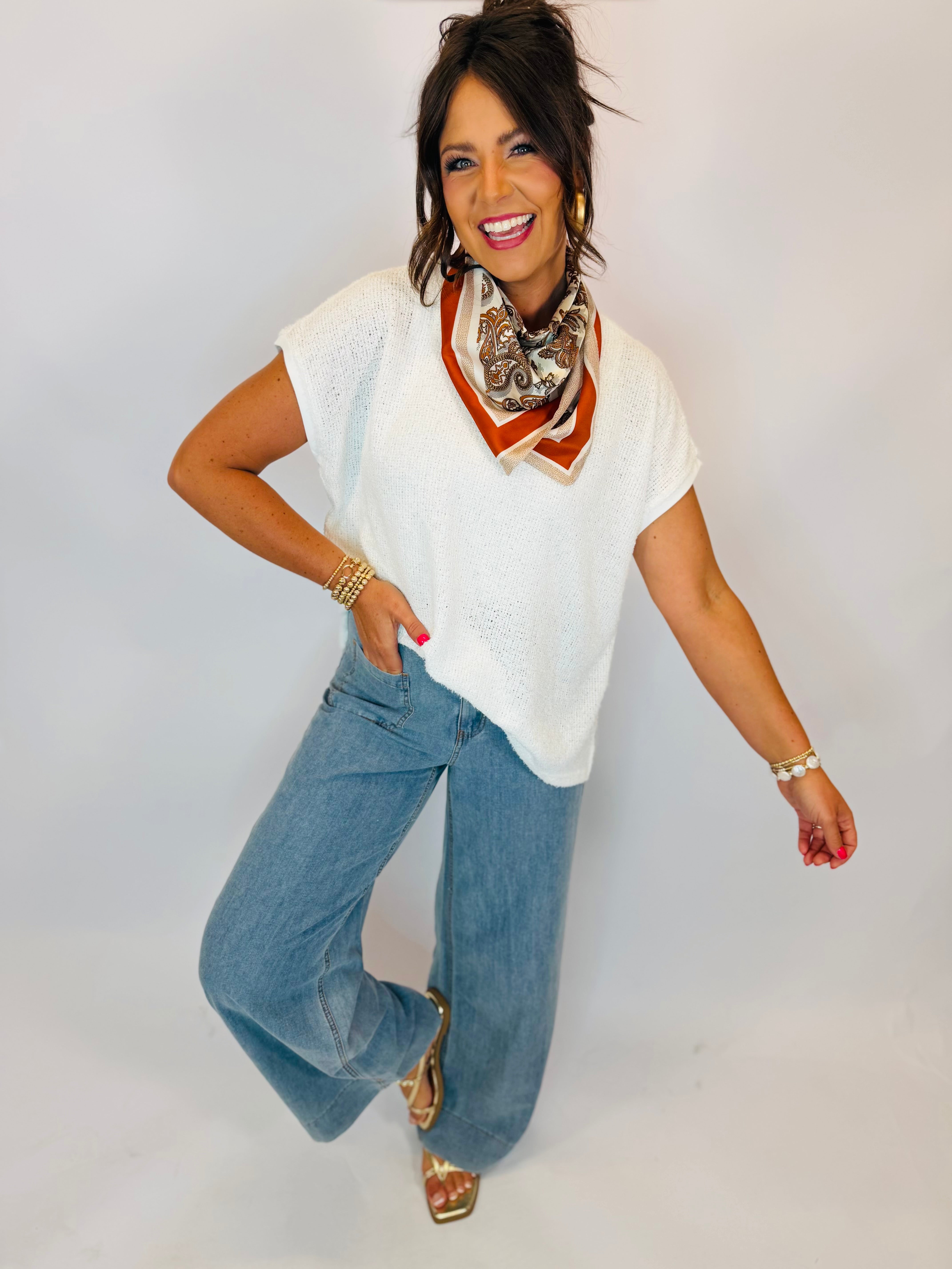 White knit sleeveless V-neck sweater top styled with wide-leg jeans and paisley neck scarf.