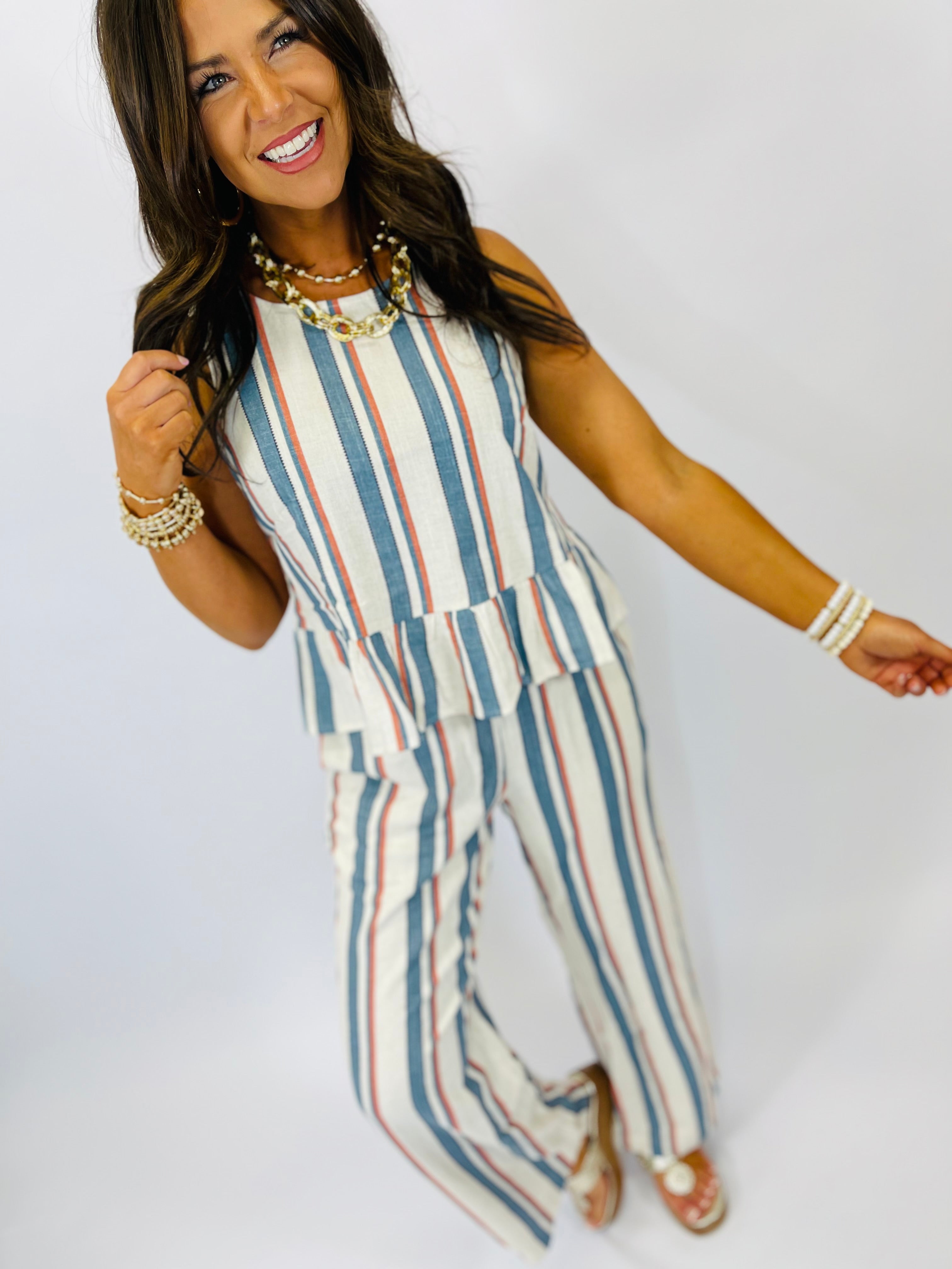 Woman wearing striped sleeveless jumpsuit in blue, white and coral with layered necklaces and bracelets.