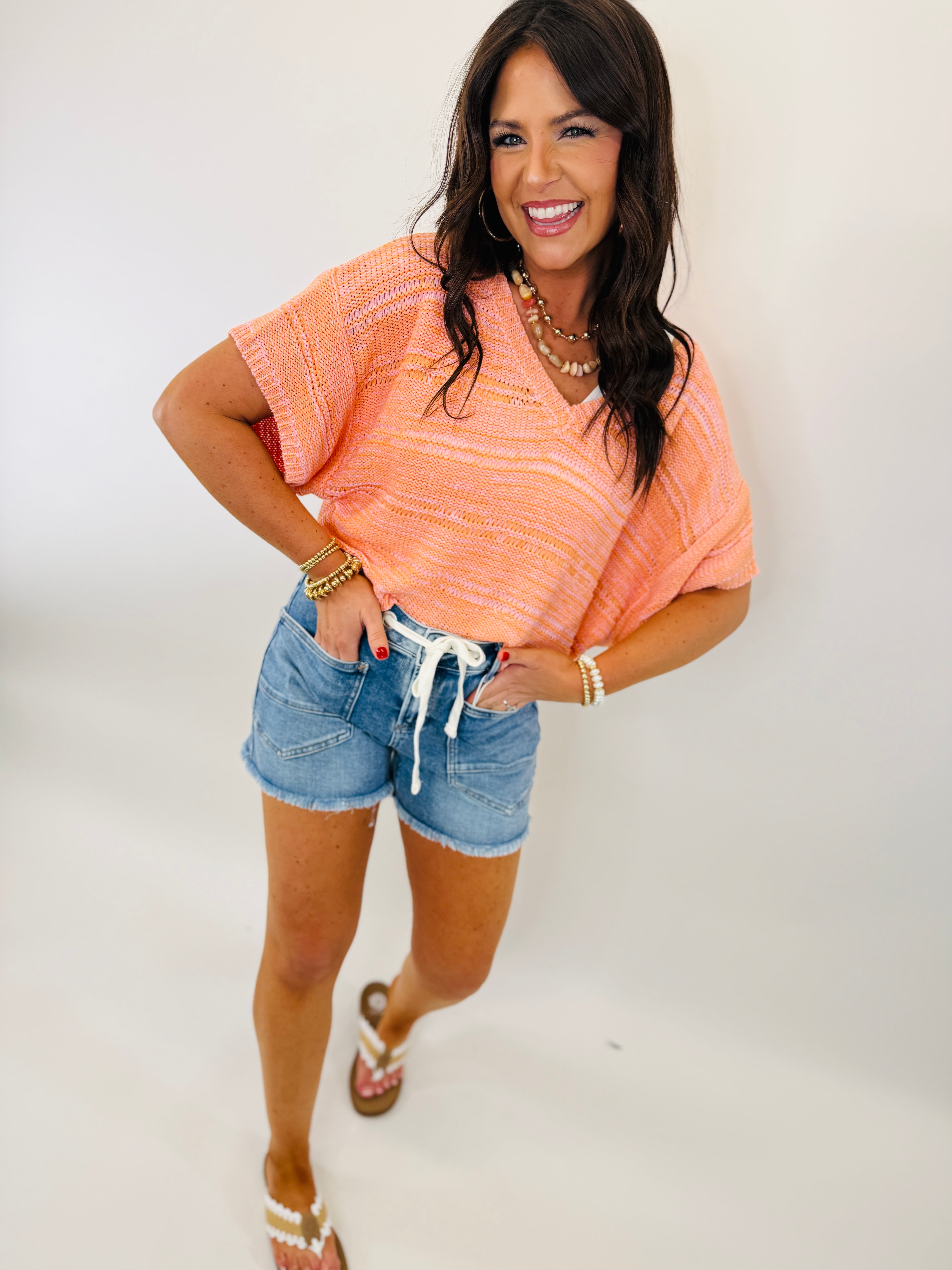 Woman wearing coral pink knit top and denim shorts with white drawstring, smiling and posing