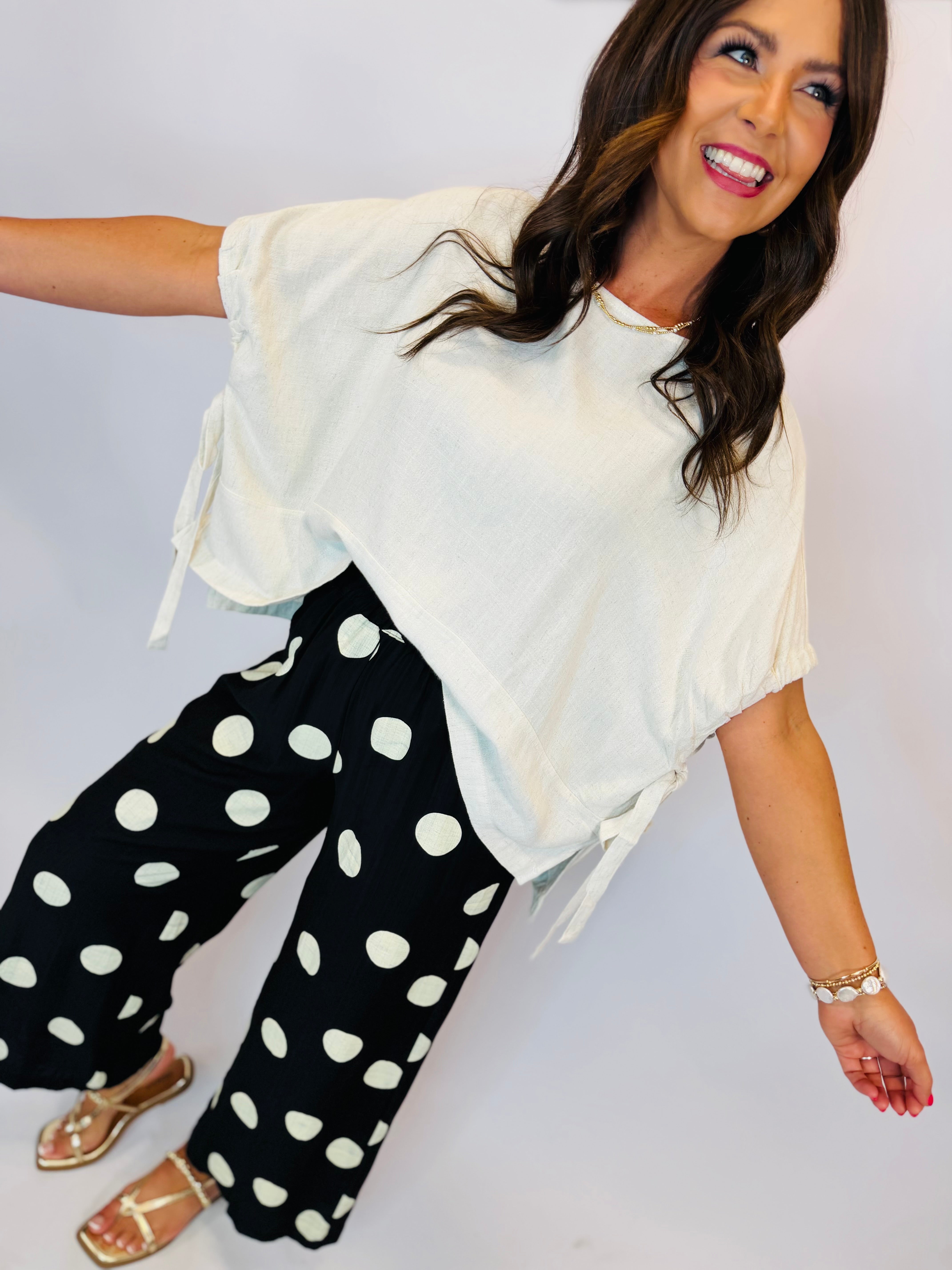 Woman wearing cream Cici Top with side ties, paired with black polka dot wide-leg pants and gold sandals.