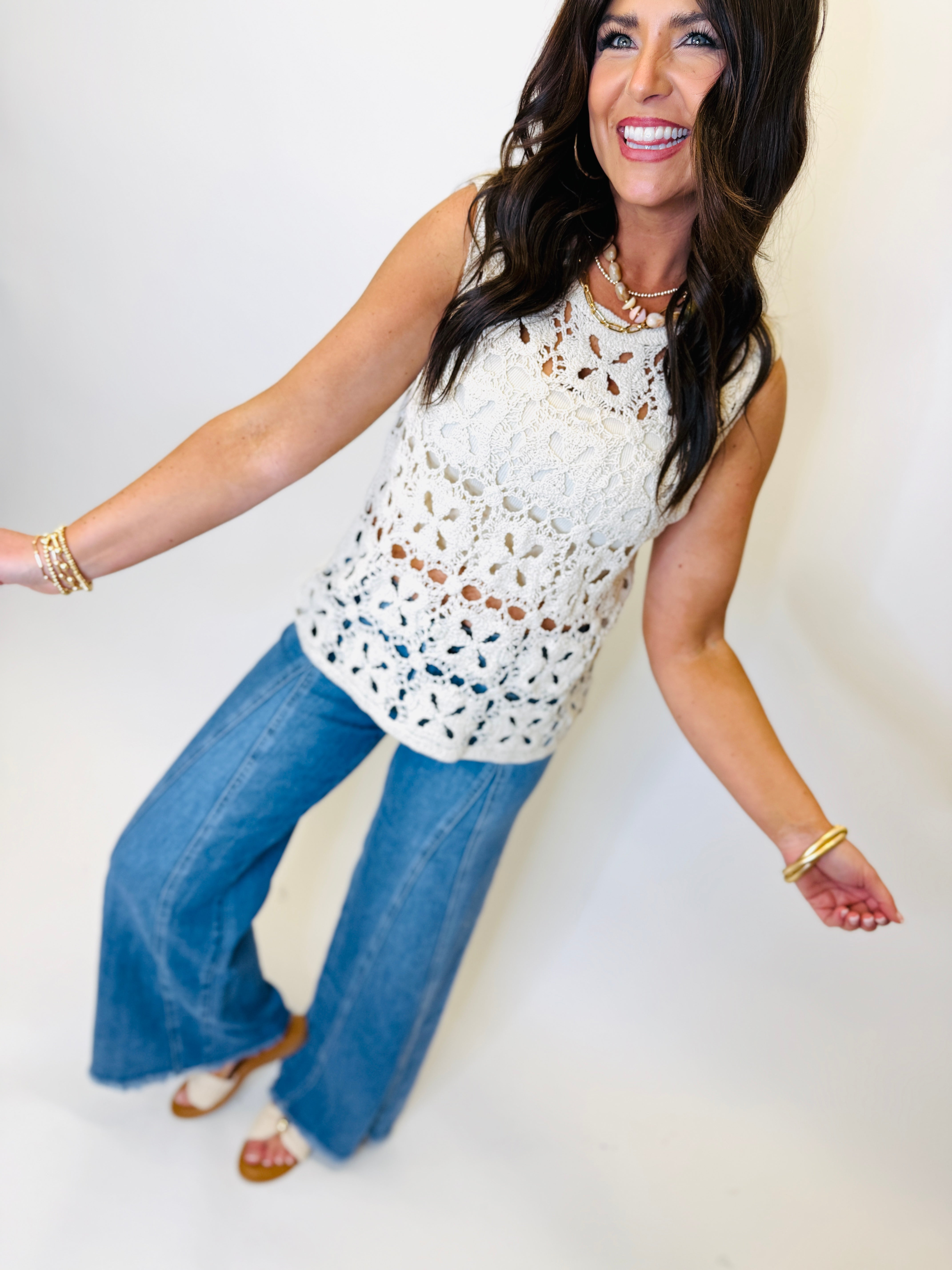 Woman wearing white crocheted sleeveless top with cutout details, paired with blue flare jeans and gold accessories.