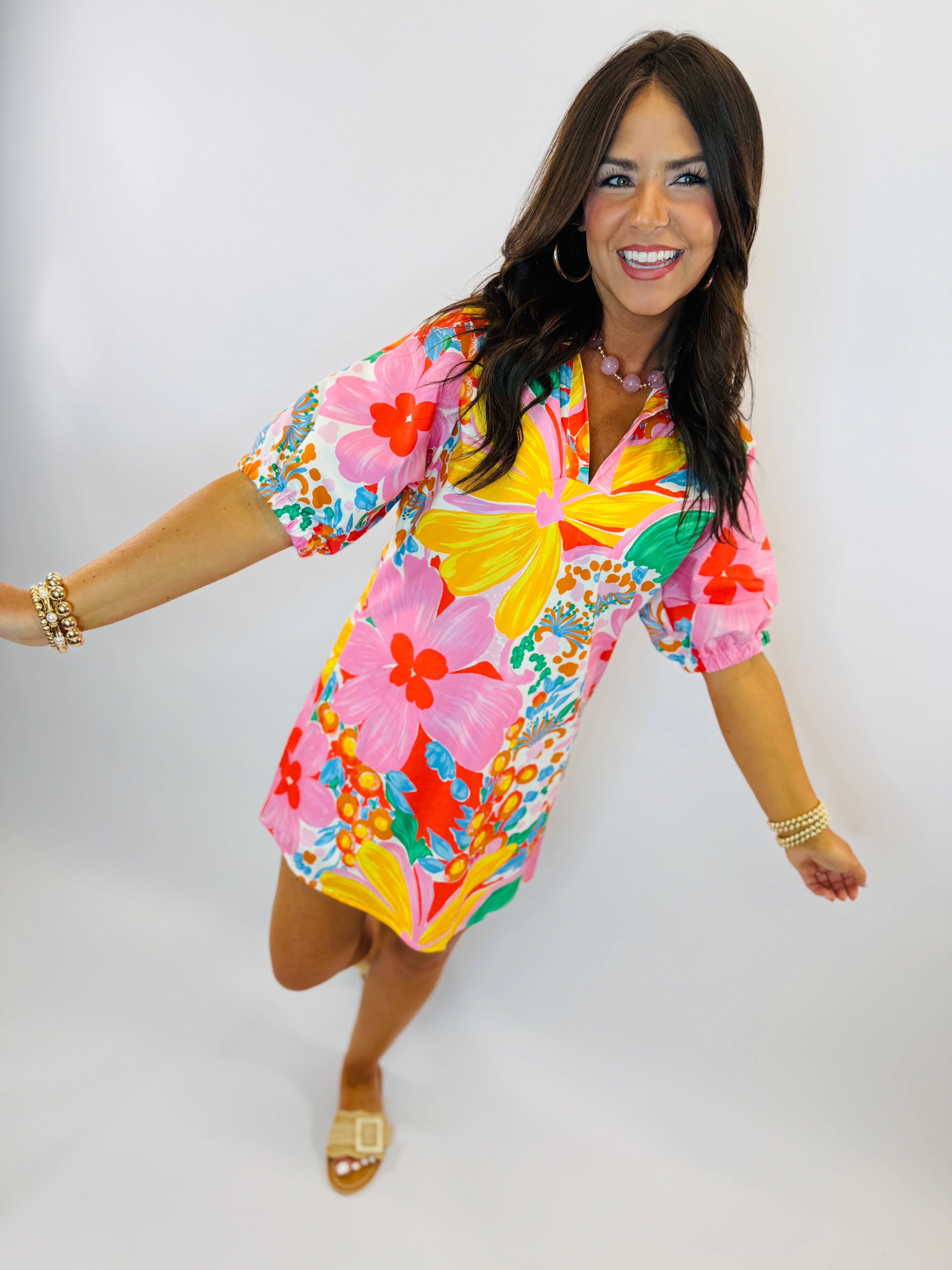 Woman wearing vibrant tropical floral print dress with short sleeves and collar, styled with gold jewelry and sandals.