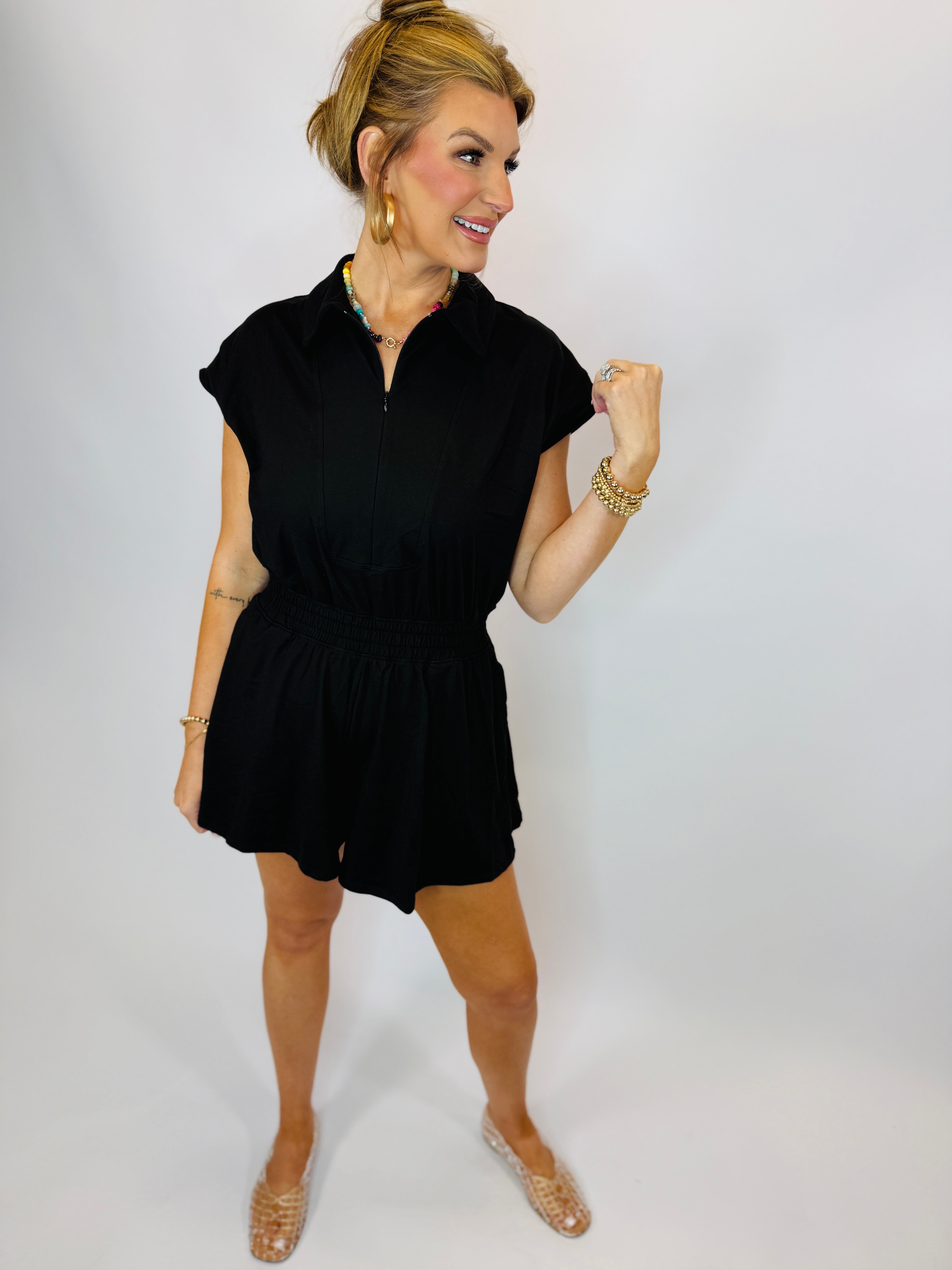 Woman wearing black short-sleeve zip-front romper with elastic waist and shorts, styled with gold jewelry and sandals.