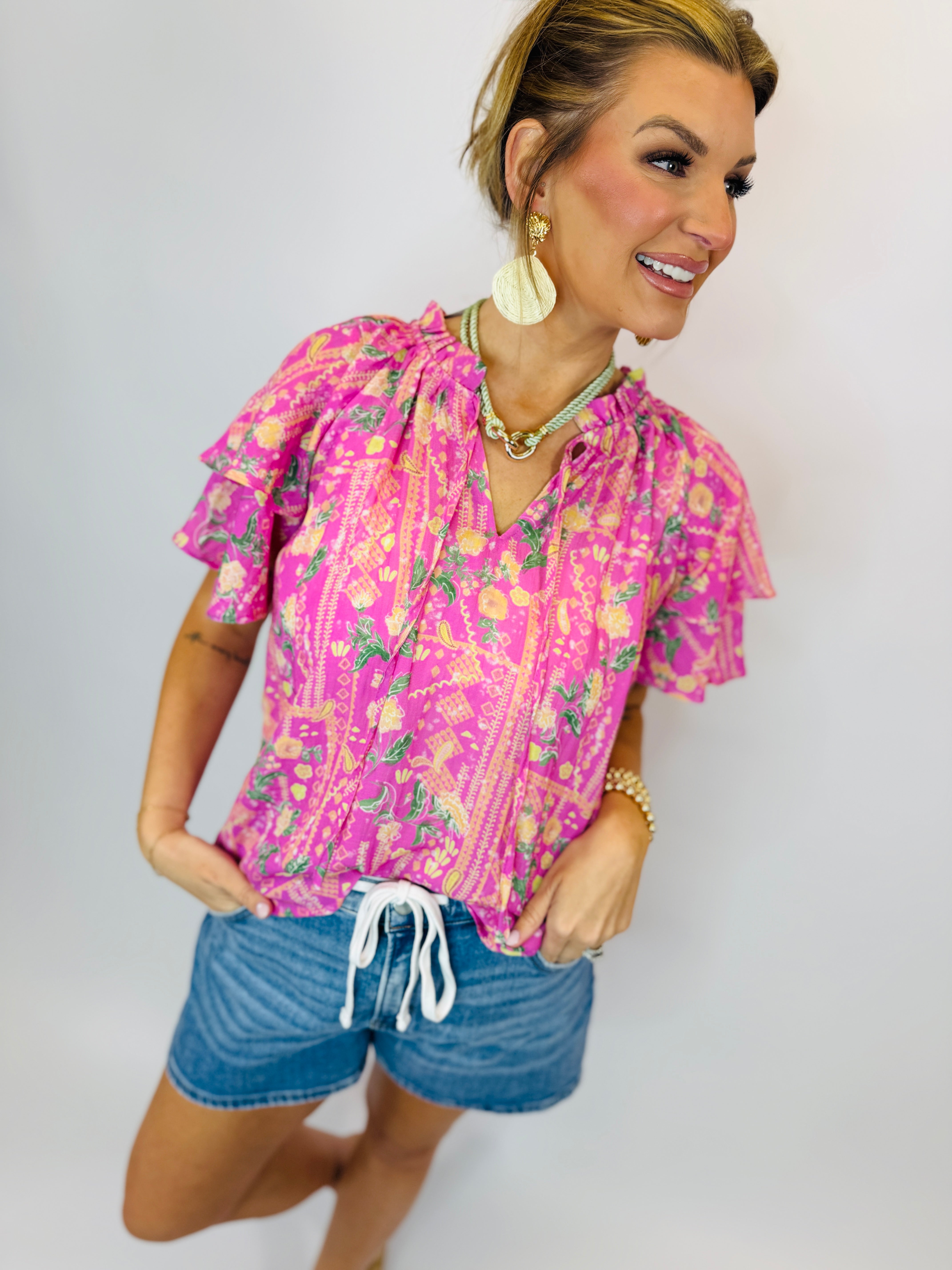 Woman wearing bright pink floral Denisse top with ruffled sleeves and drawstring neckline, paired with denim shorts.