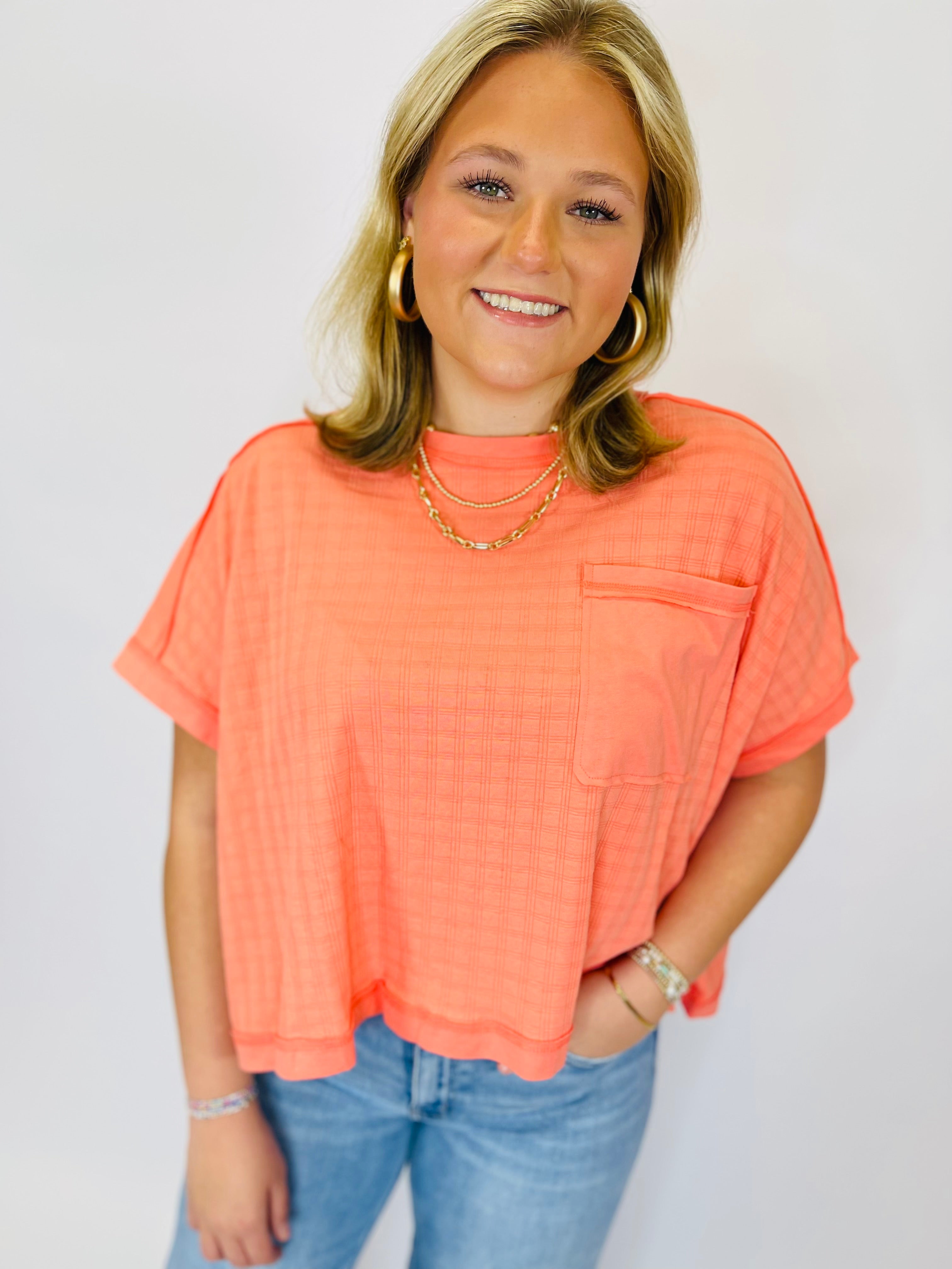 Woman wearing coral oversized short-sleeve top with chest pocket and gold layered necklaces, paired with blue jeans.