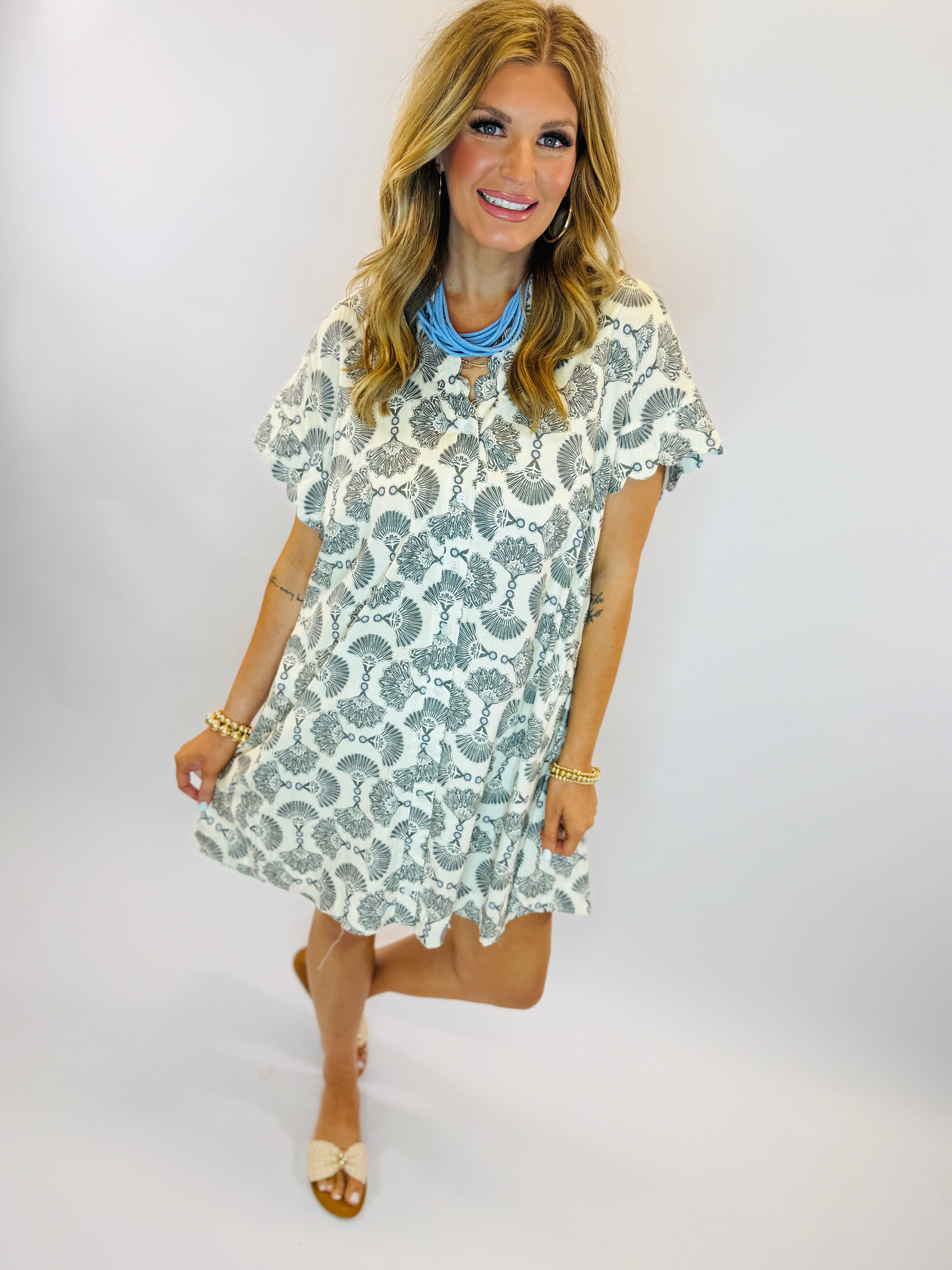 Woman wearing white short-sleeve dress with blue seashell pattern, blue necklace, and gold bracelets.
