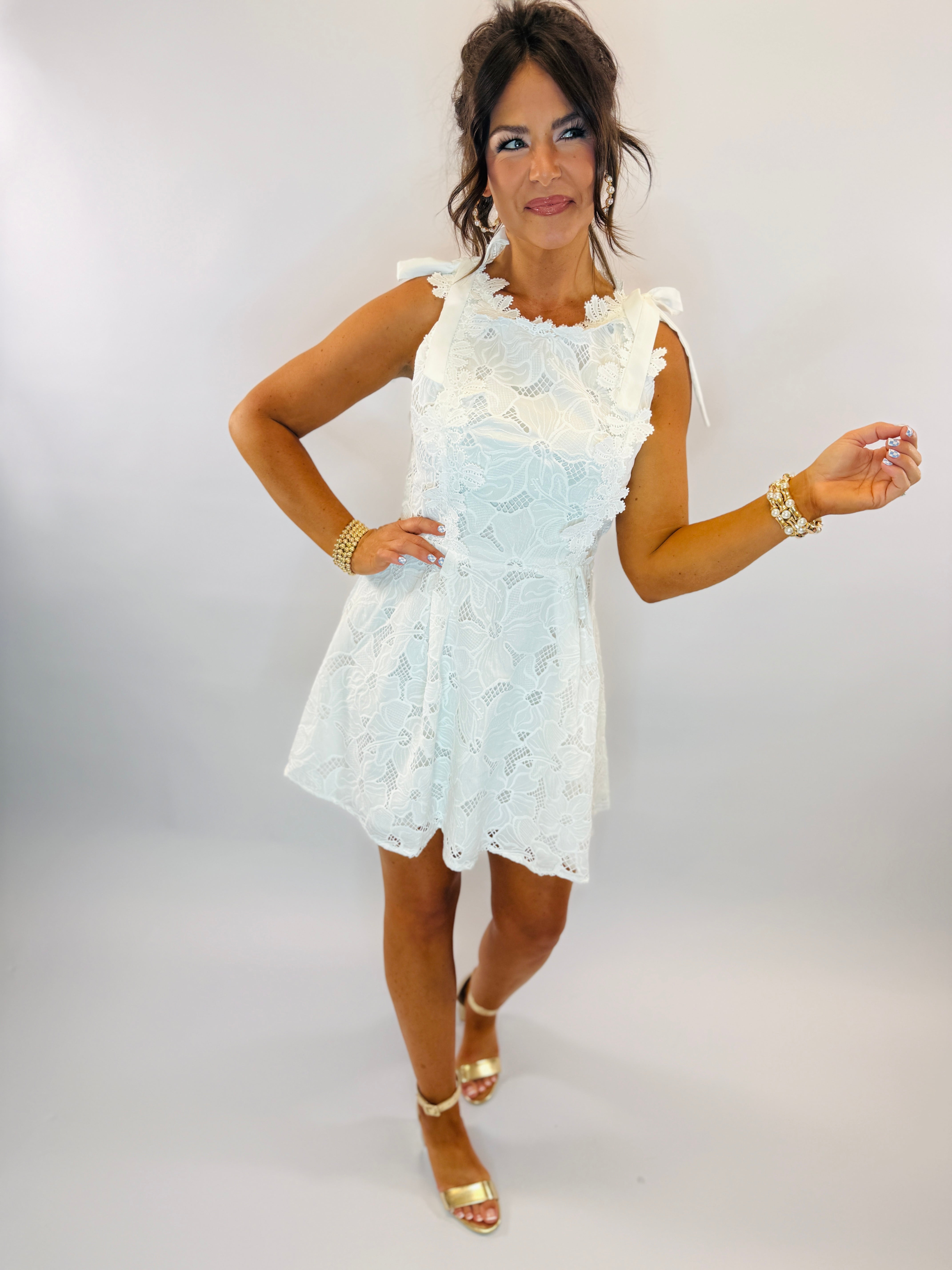 Woman modeling white lace sleeveless mini dress with scalloped neckline and gold sandals against white background.