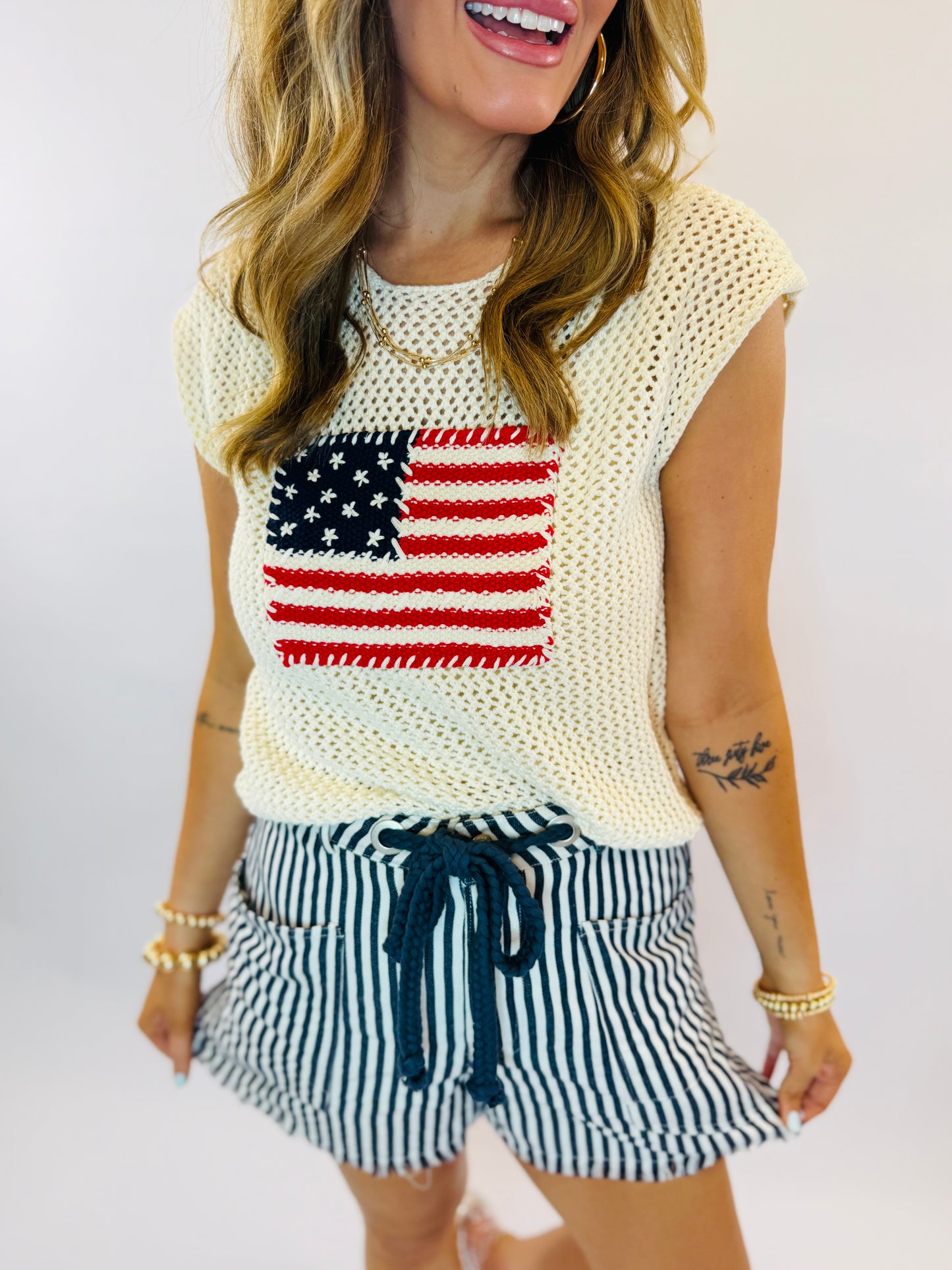 Woman wearing cream knit American flag sweater and navy striped drawstring shorts with ruffled hem.