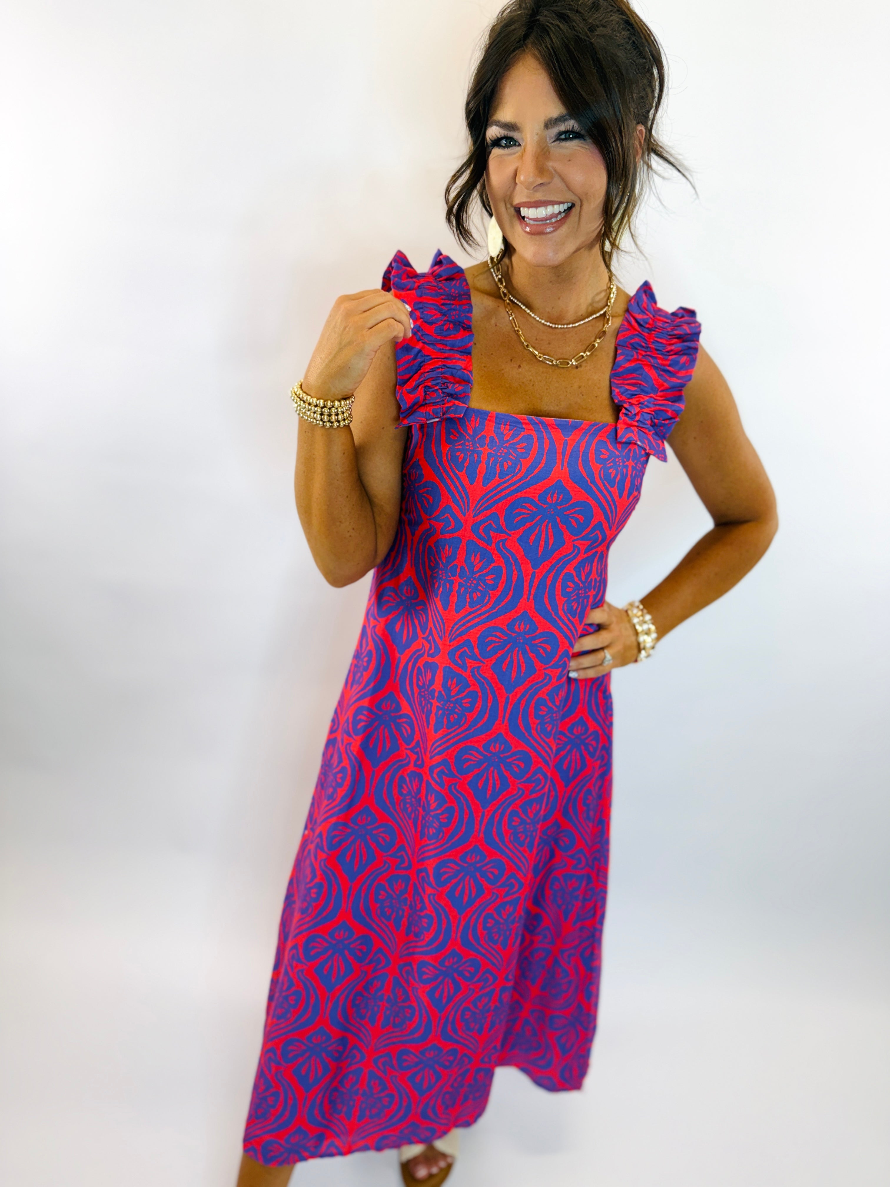 Woman wearing red and blue patterned Ciara dress with ruffle straps and square neckline, accessorized with gold jewelry.