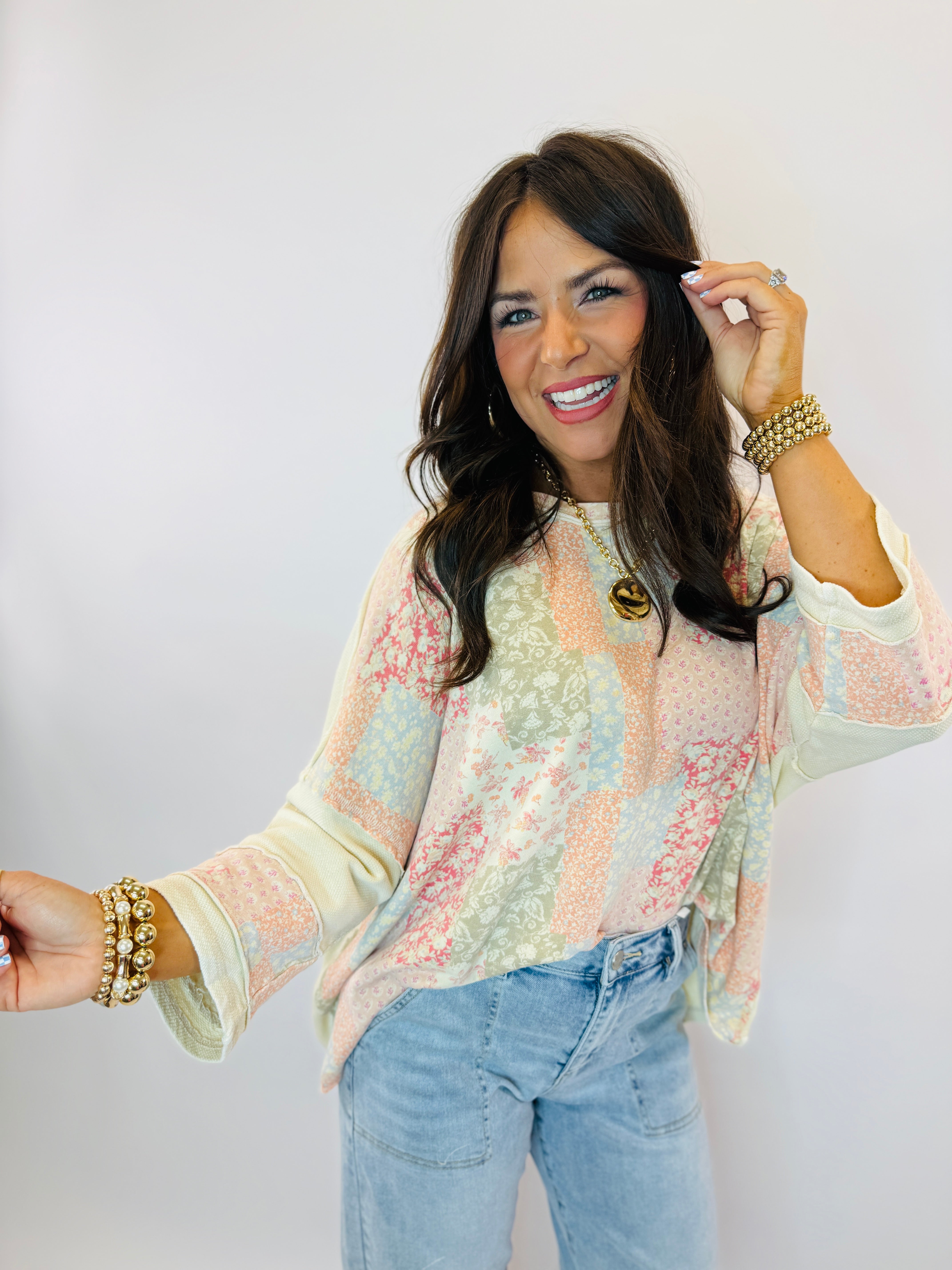 Woman wearing pastel patchwork floral Ellie top with long sleeves, paired with light blue jeans and gold jewelry.