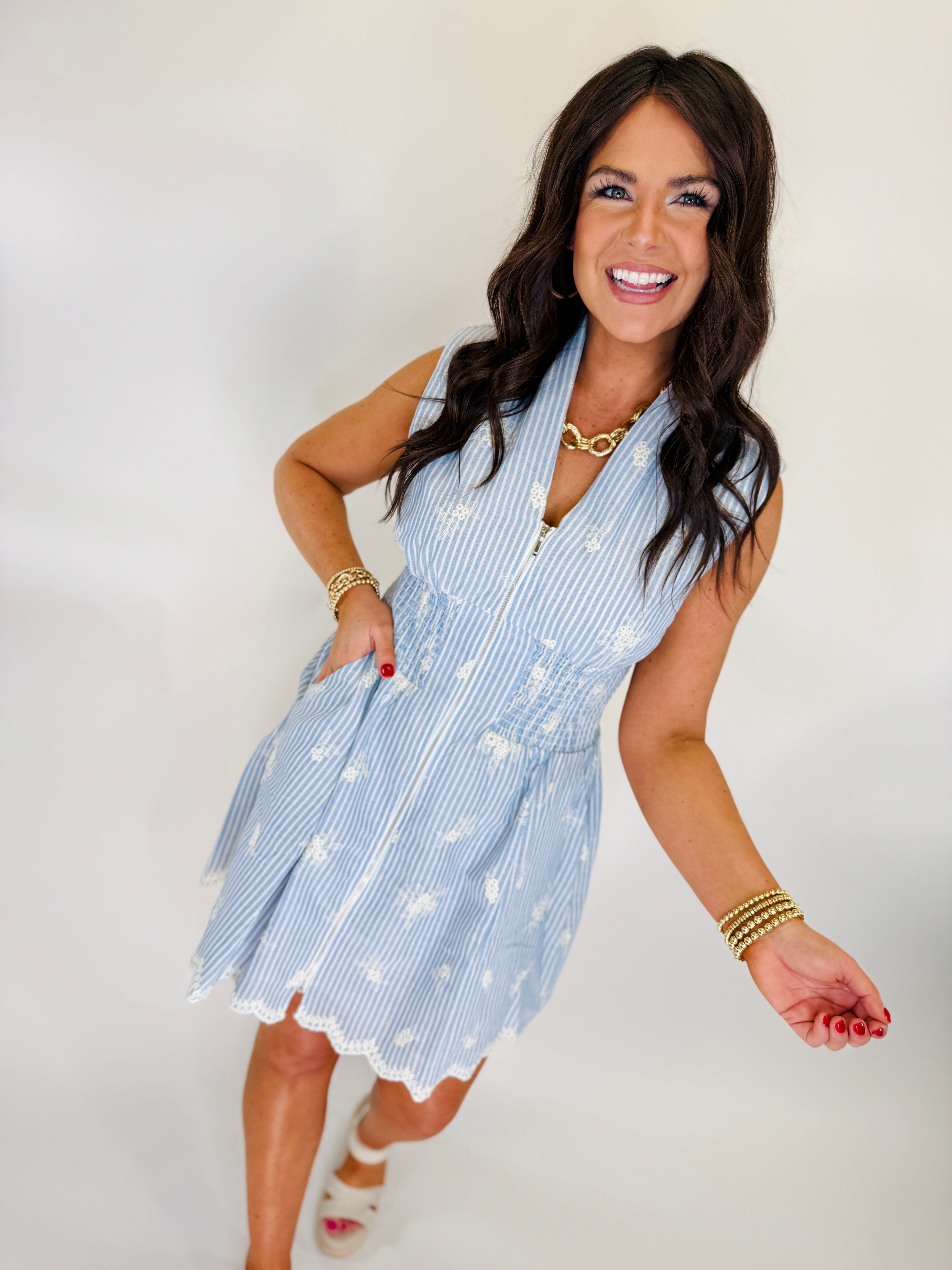 Woman wearing light blue striped sleeveless dress with white floral embroidery and gold jewelry accessories.