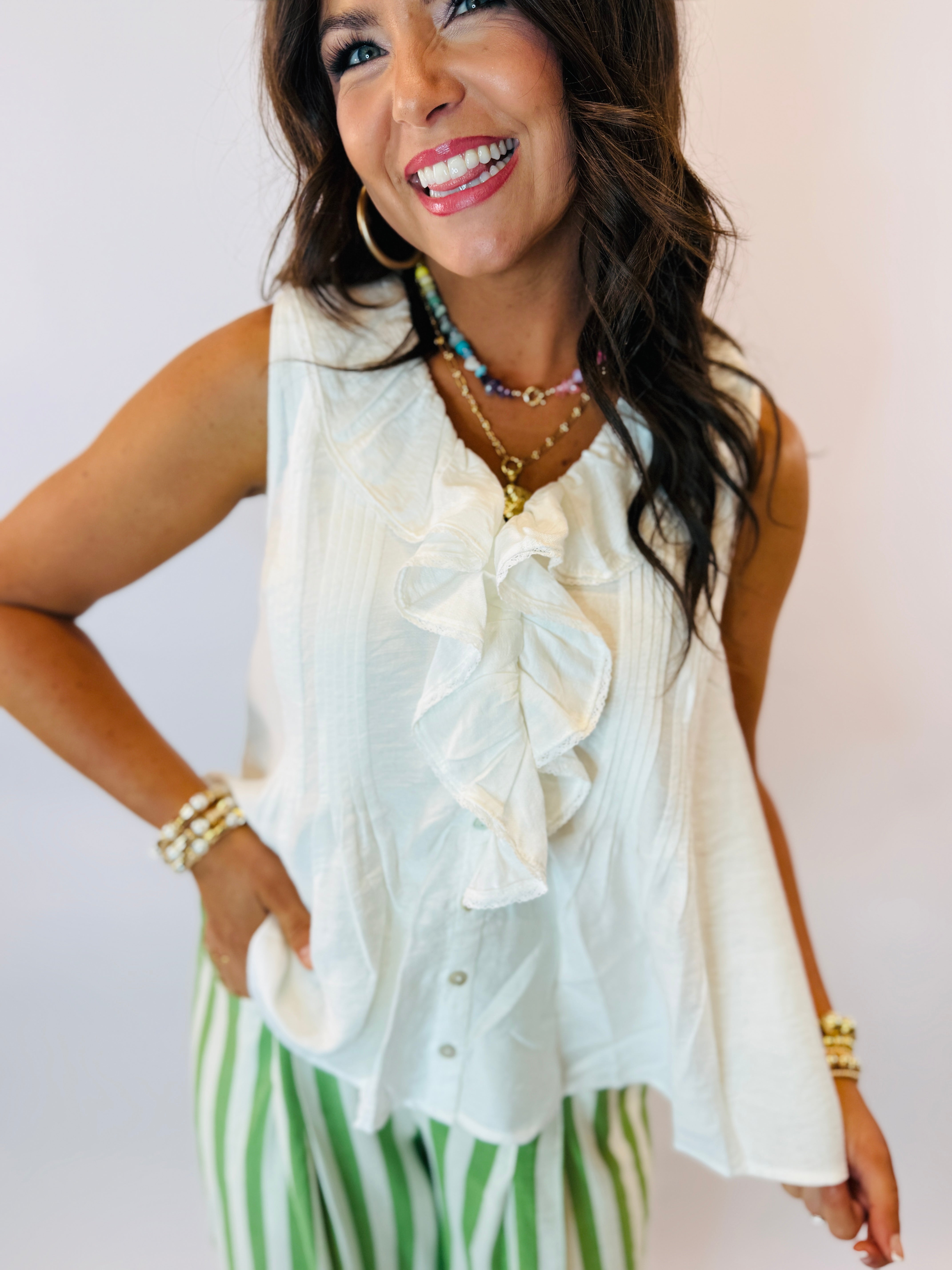 Woman wearing white sleeveless ruffled blouse with green and white striped pants, styled with layered jewelry.