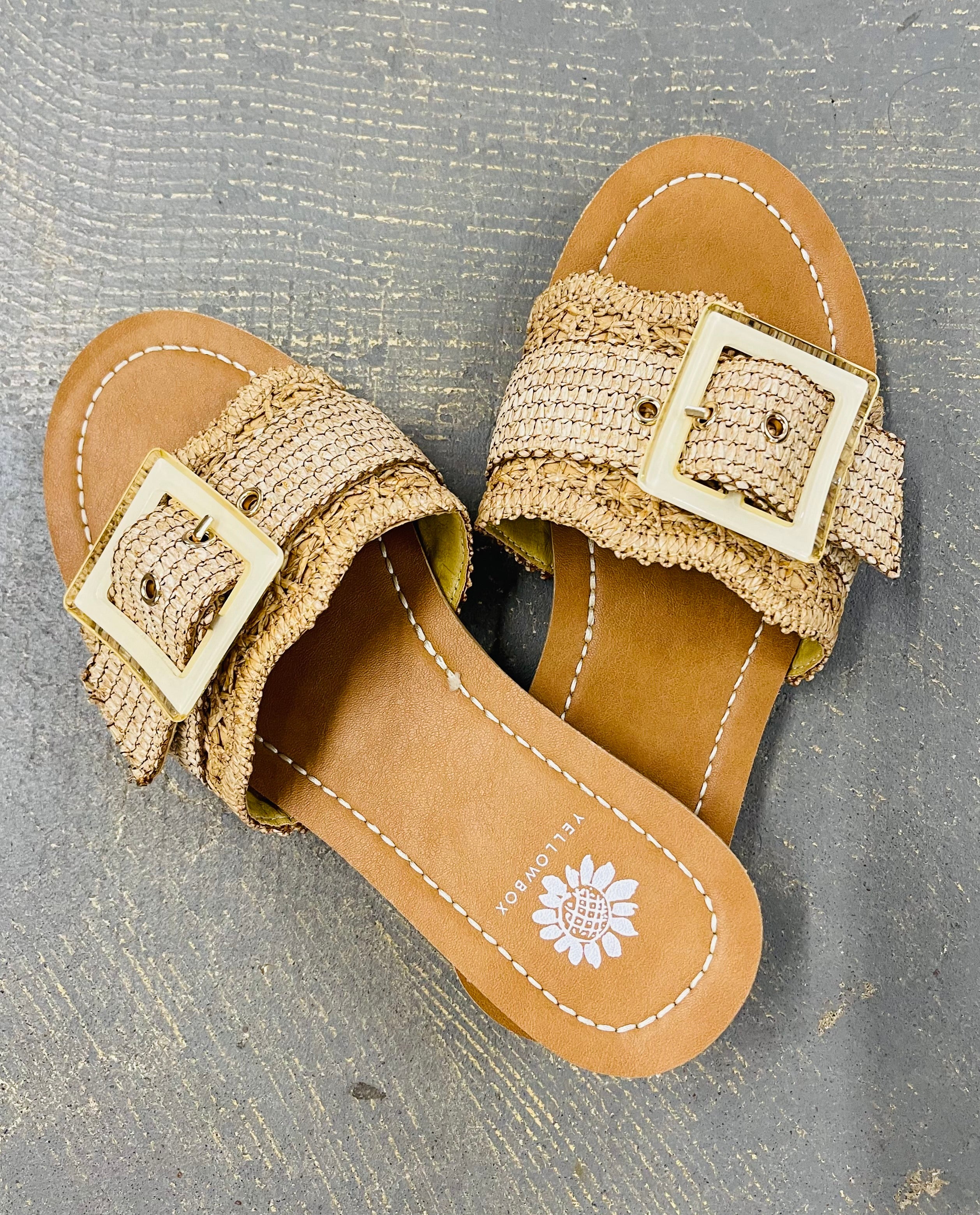 Tan woven sandals with gold square buckles and white stitching detail on wooden surface.