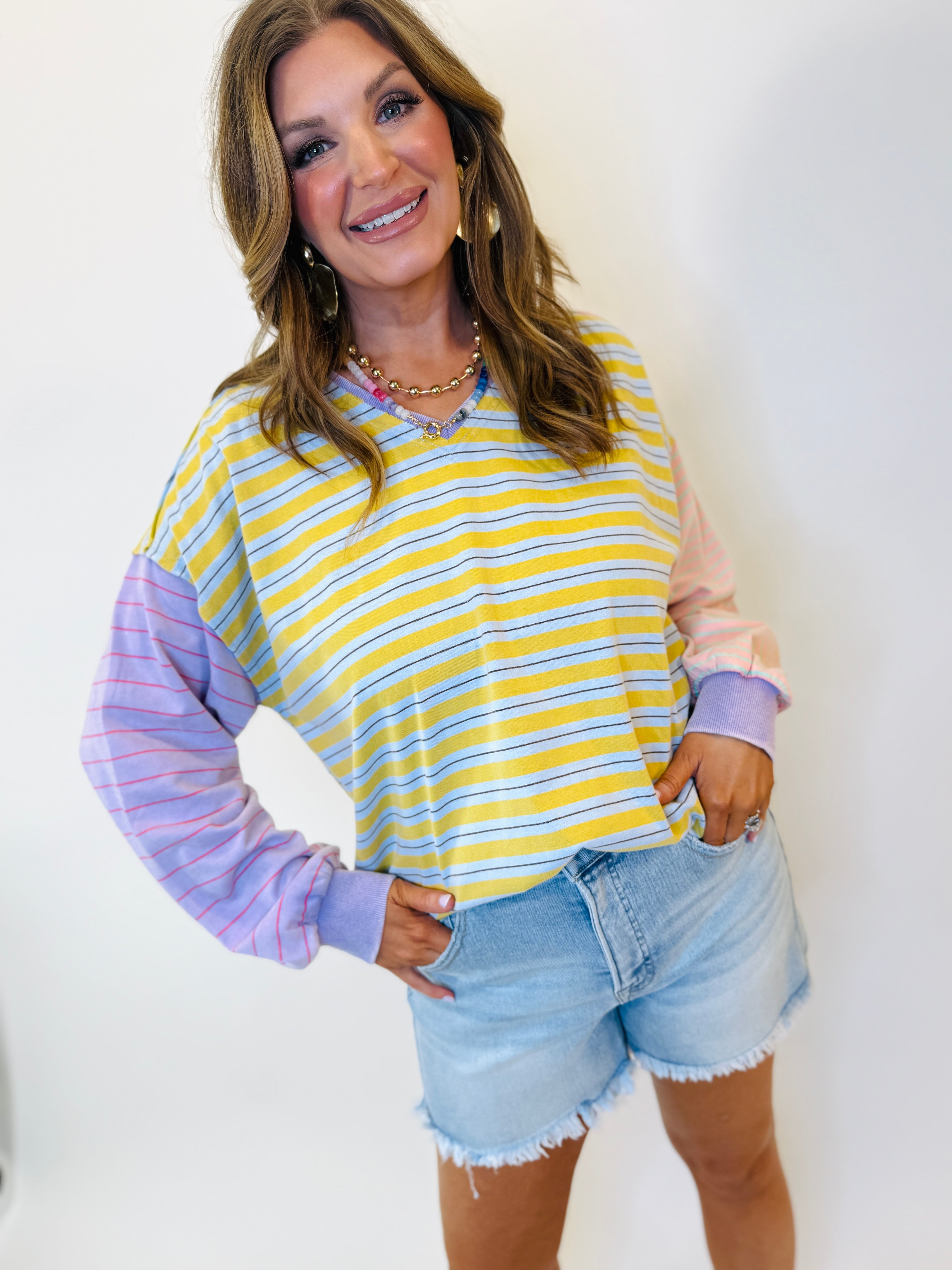 Woman wearing colorful striped Emmie pullover with yellow, blue, and purple stripes paired with denim shorts.
