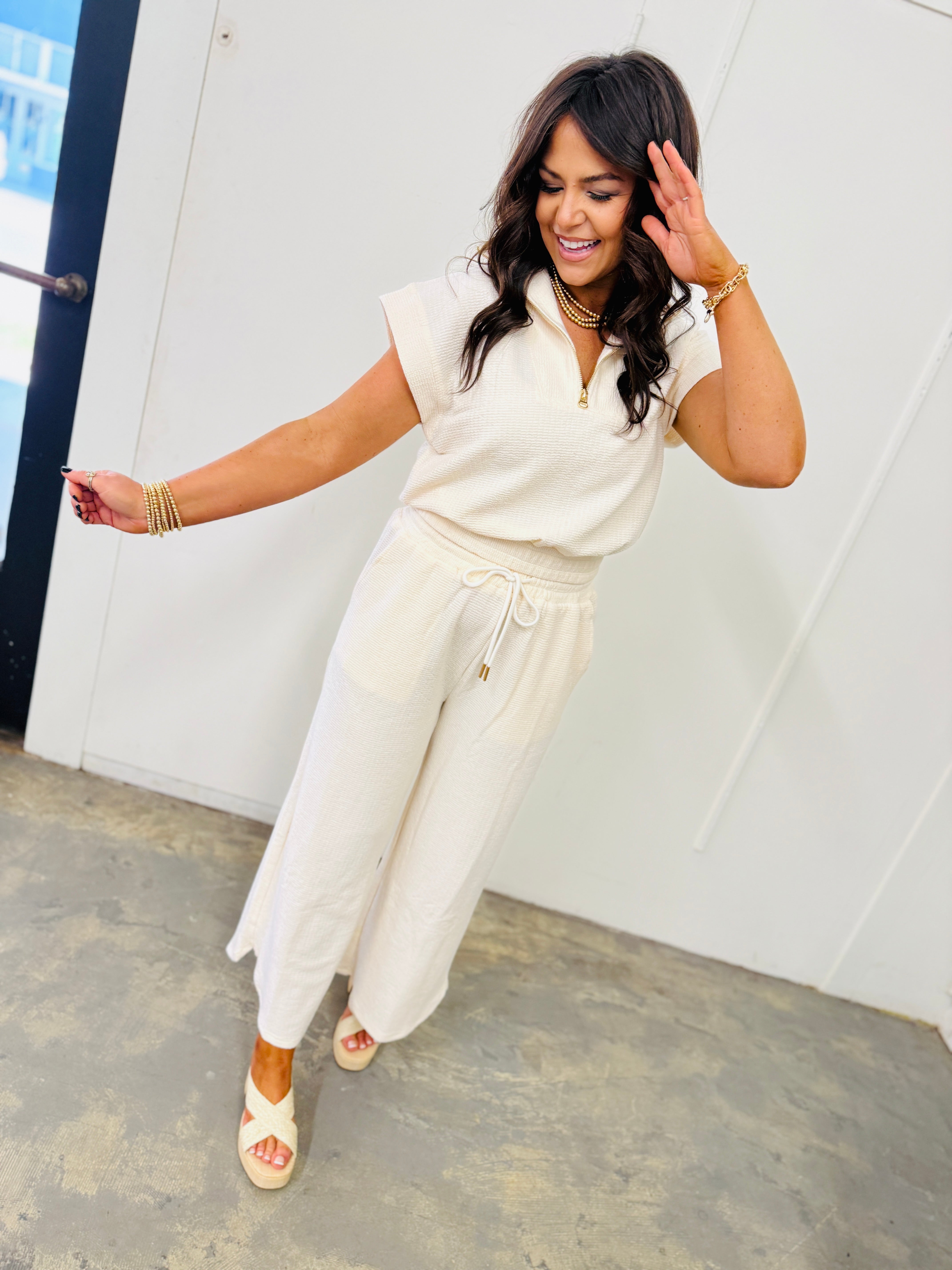 Woman wearing cream-colored sleeveless knit top with drawstring waist, styled with gold jewelry and platform sandals.