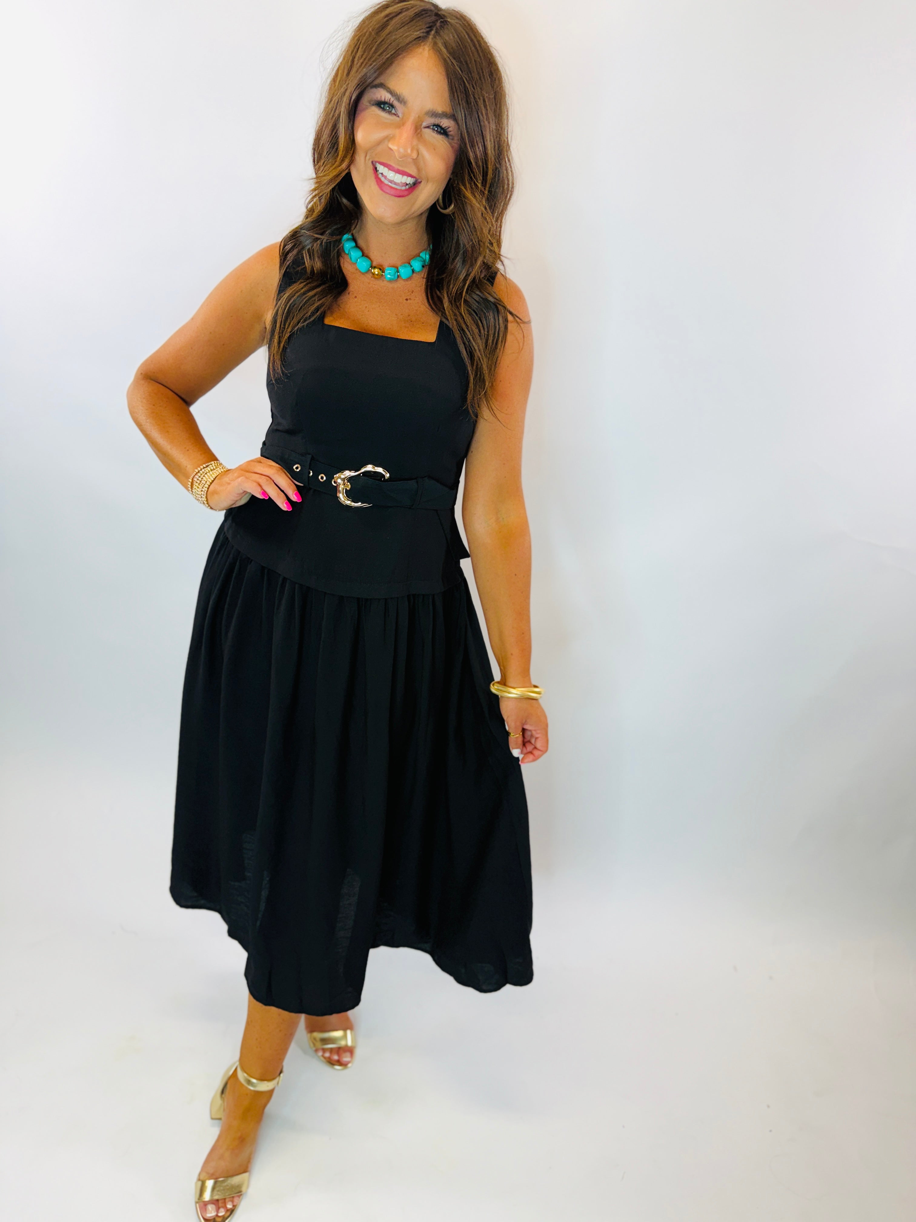 Woman wearing black sleeveless midi dress with gold belt buckle, styled with turquoise necklace and gold heels.