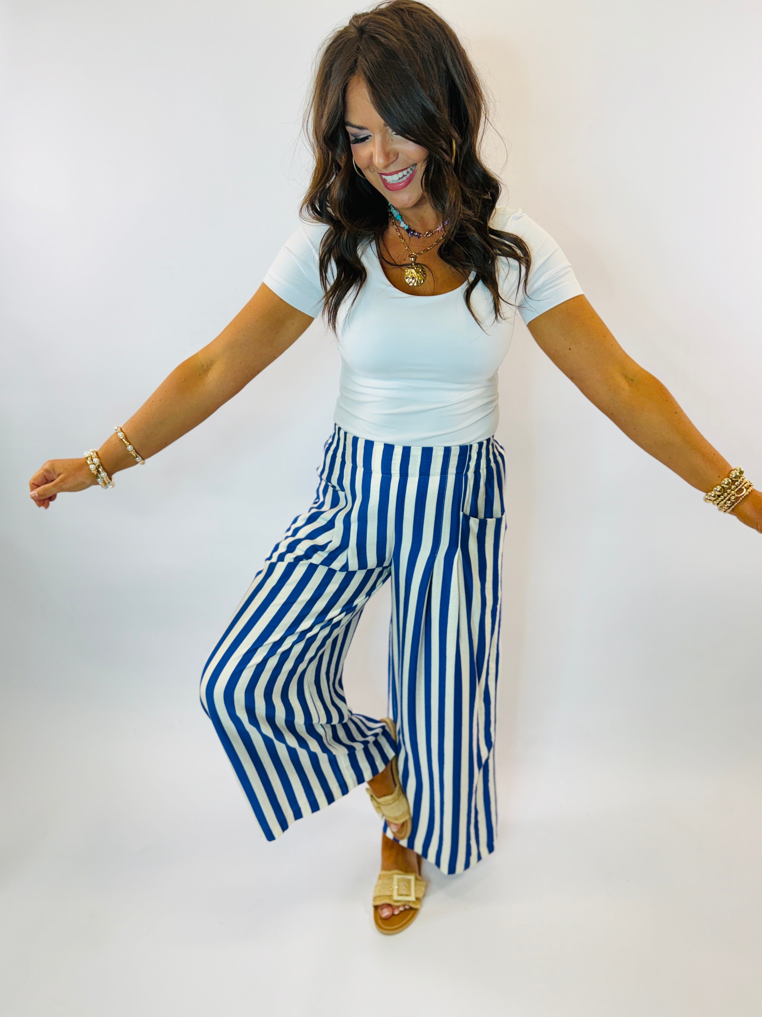Woman wearing white top and blue and white striped wide-leg pants with gold accessories and sandals.