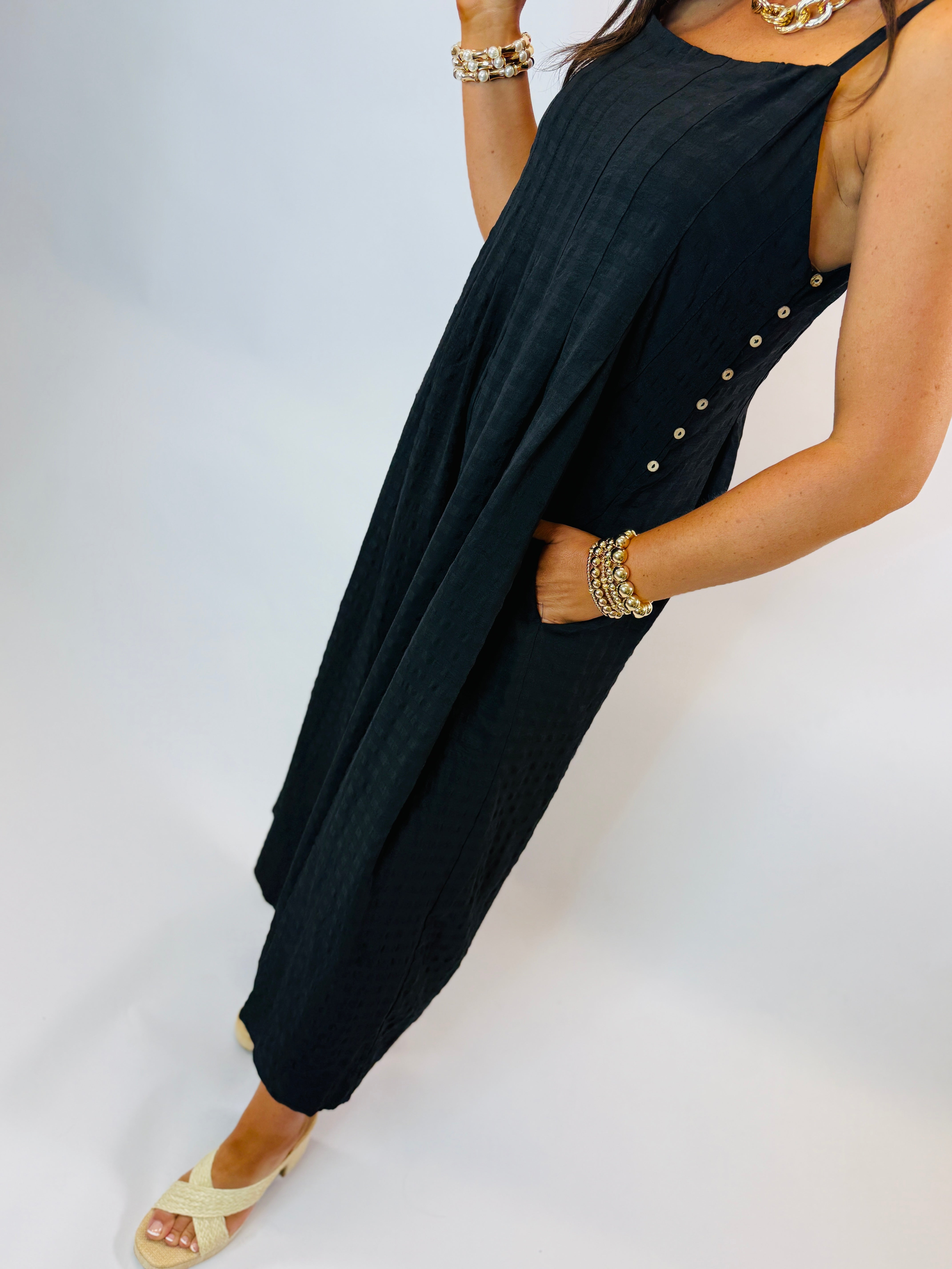 Navy blue sleeveless pleated jumpsuit with side button details and pockets, styled with gold jewelry and platform sandals.