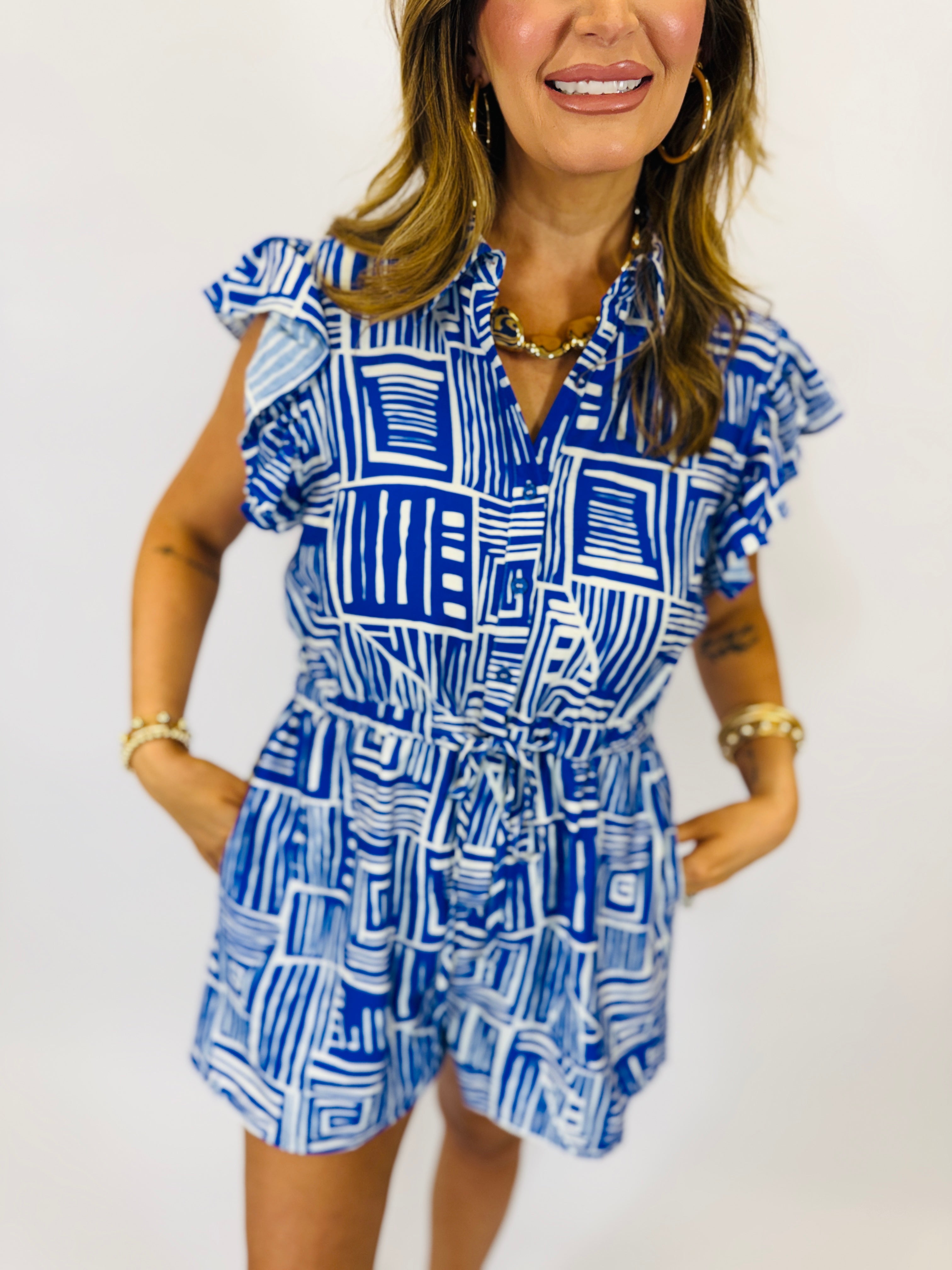 Woman wearing blue and white geometric patterned romper with ruffled sleeves and tie waist belt.