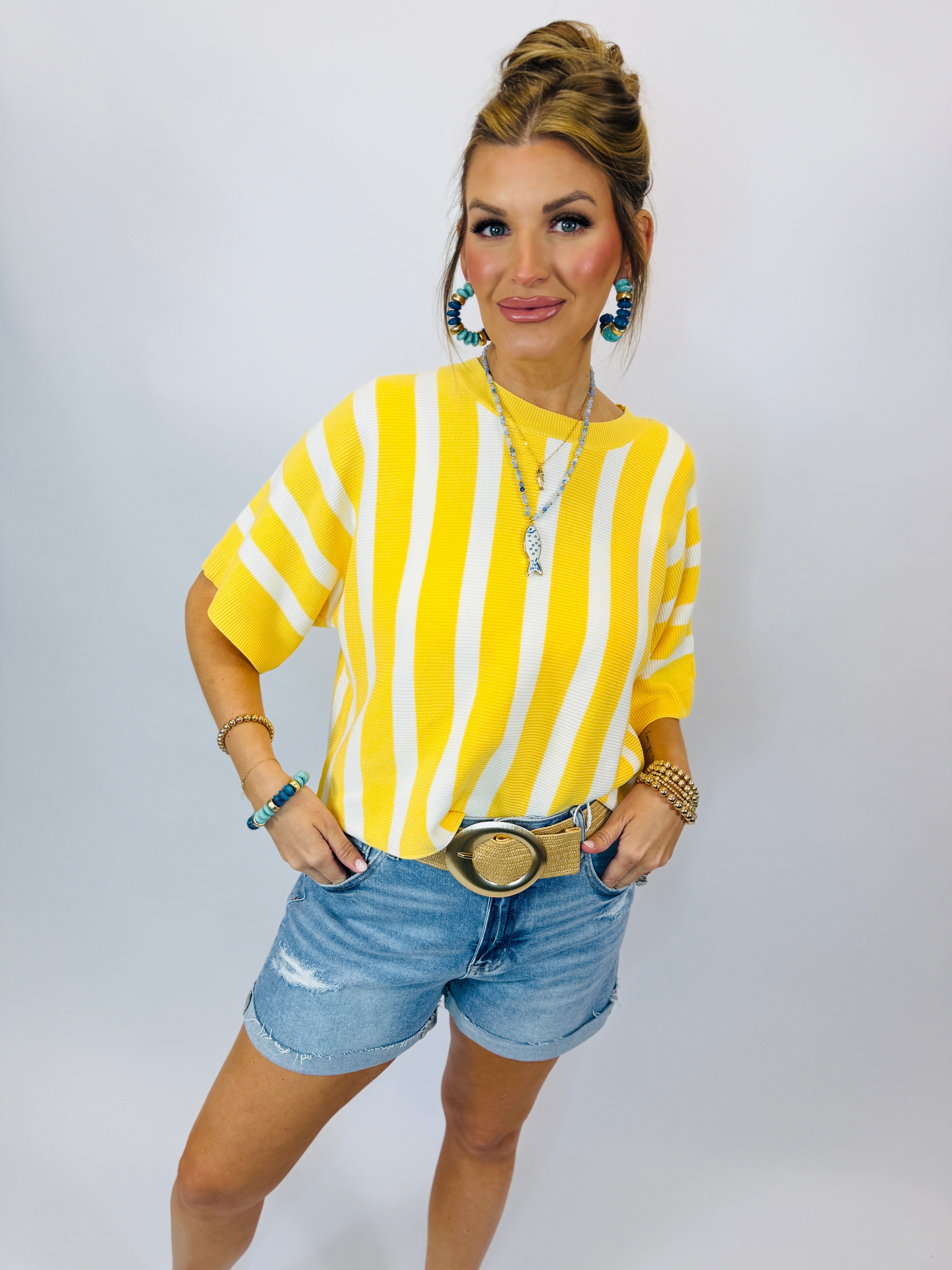 Woman wearing yellow and white striped short-sleeve sweater with denim shorts and layered jewelry accessories.
