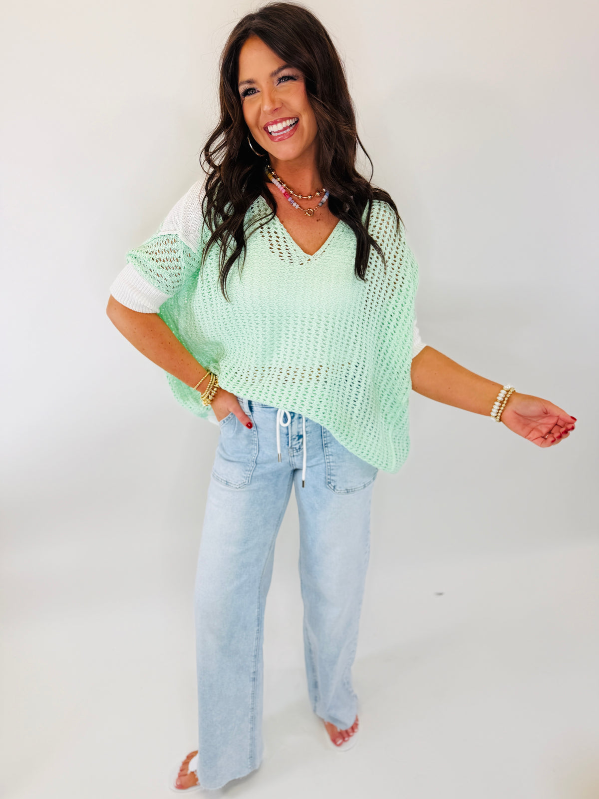 Woman wearing mint green knit sweater with white sleeves and light blue jeans, smiling against white background.