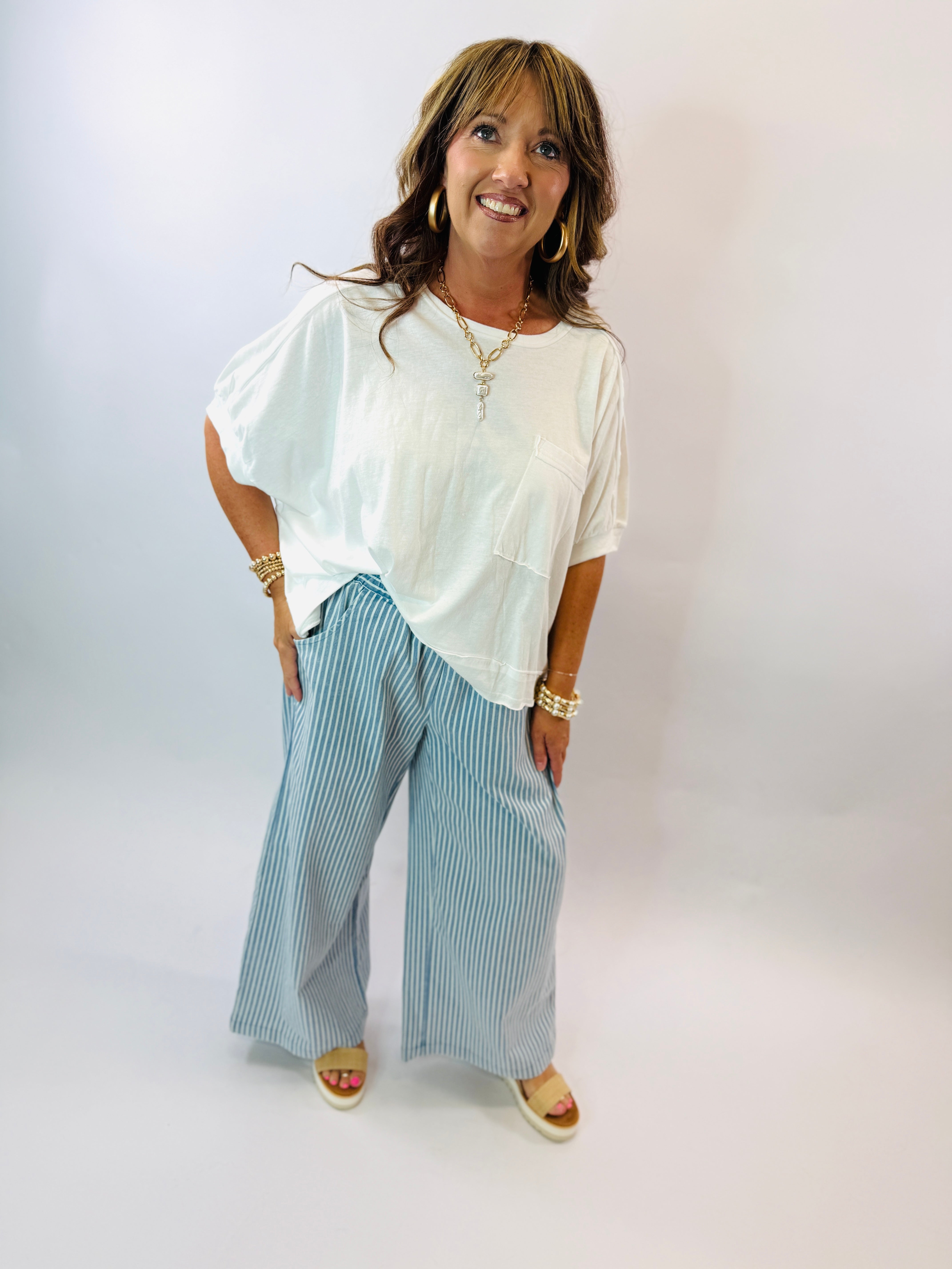 Woman wearing white oversized top and blue striped wide-leg pants with gold accessories and sandals.
