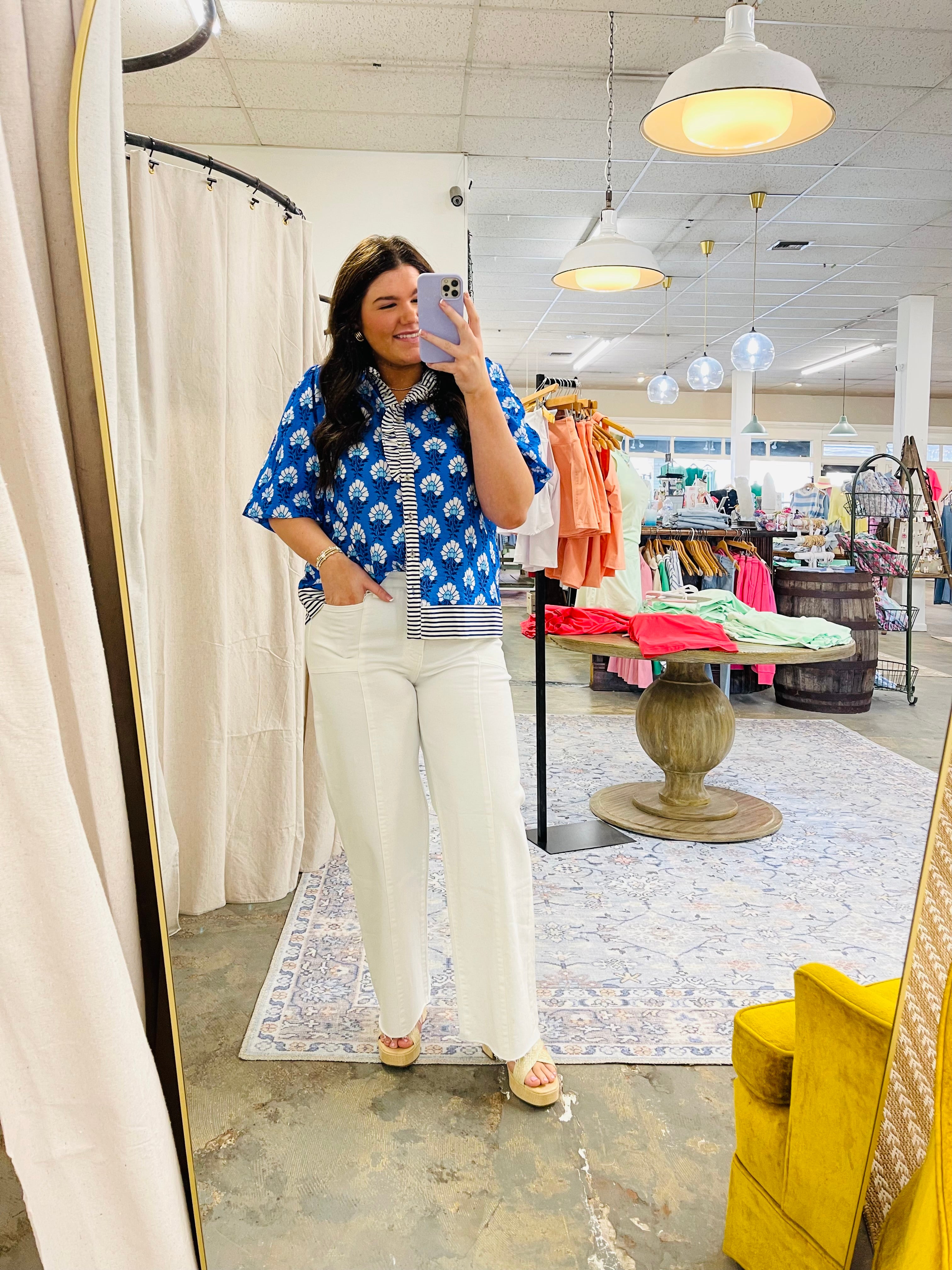 Woman wearing blue floral Sami Top with striped undershirt and white pants, taking mirror selfie in clothing store