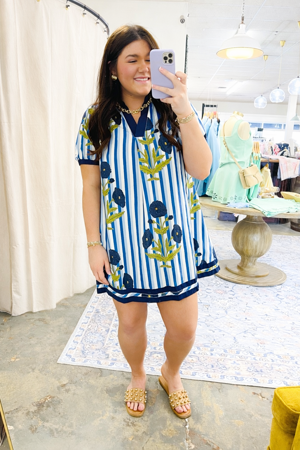 Woman wearing blue and white striped dress with floral pattern taking mirror selfie in boutique store